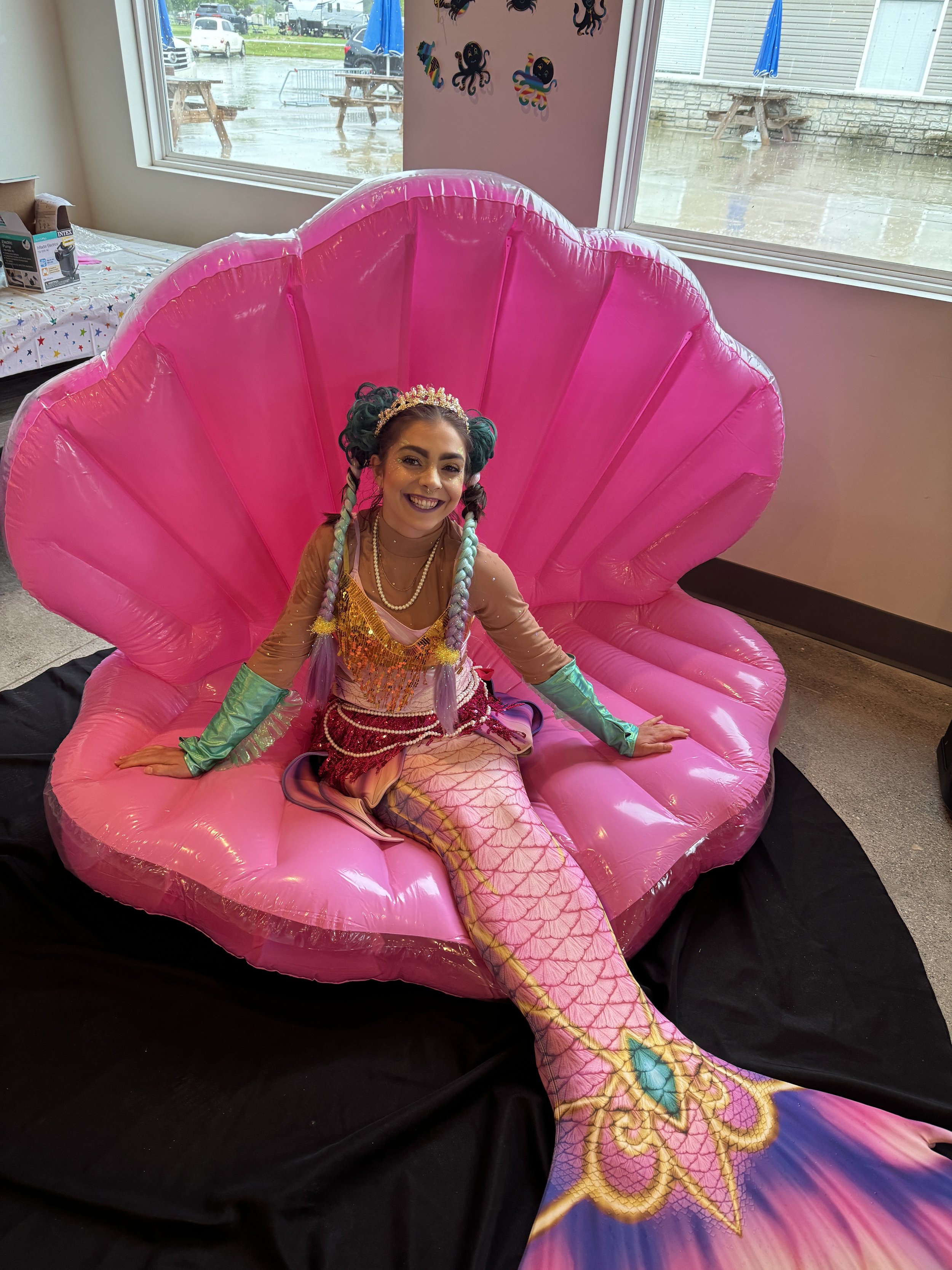 Young woman dressed as a mermaid sitting inside a large pink clam shell prop with an ocean-themed background.