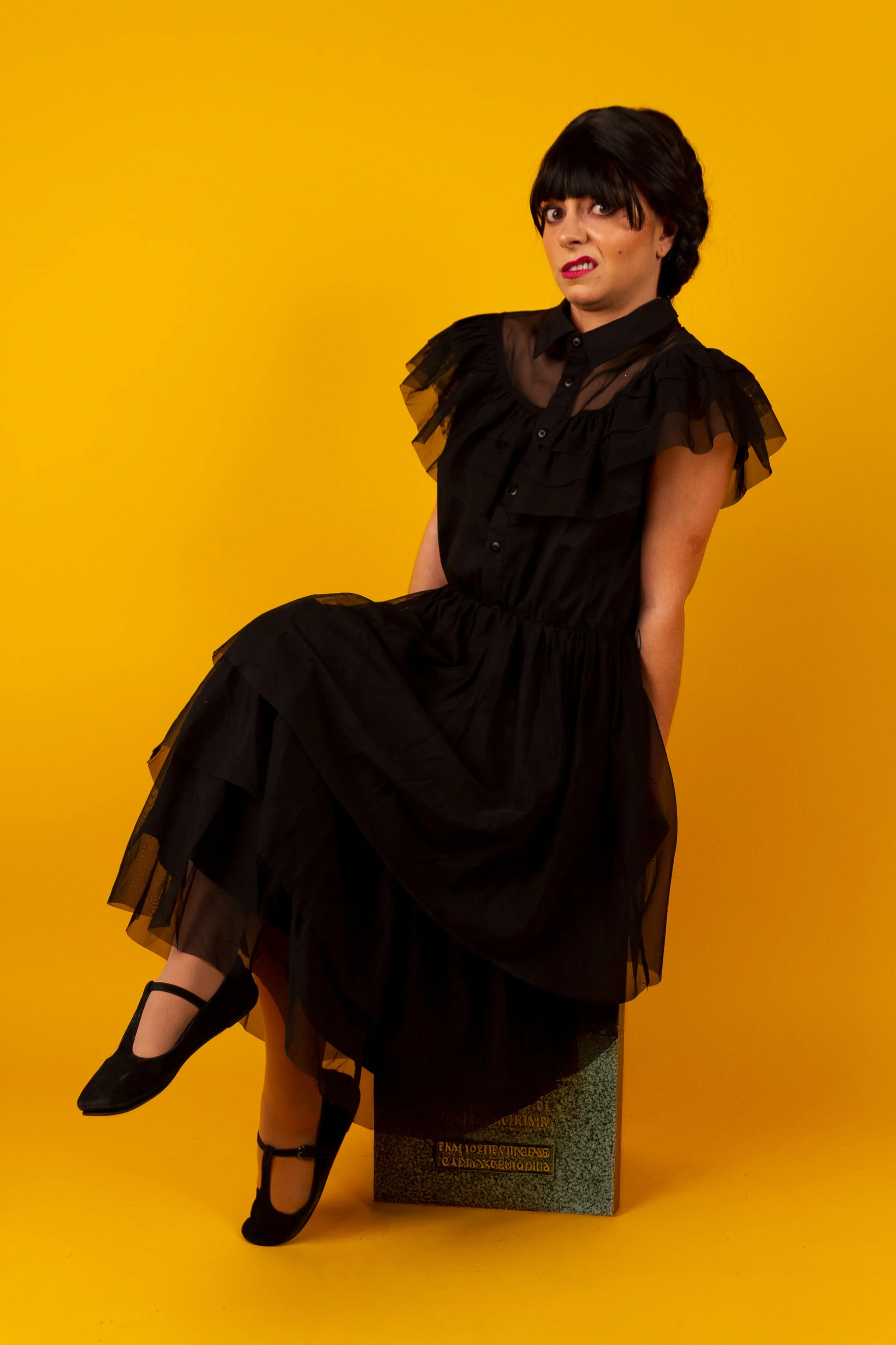 A woman with short dark hair and bangs, wearing a black tulle dress, sitting on a rectangular block against a yellow background. She has an expressive facial expression and is looking at the camera.