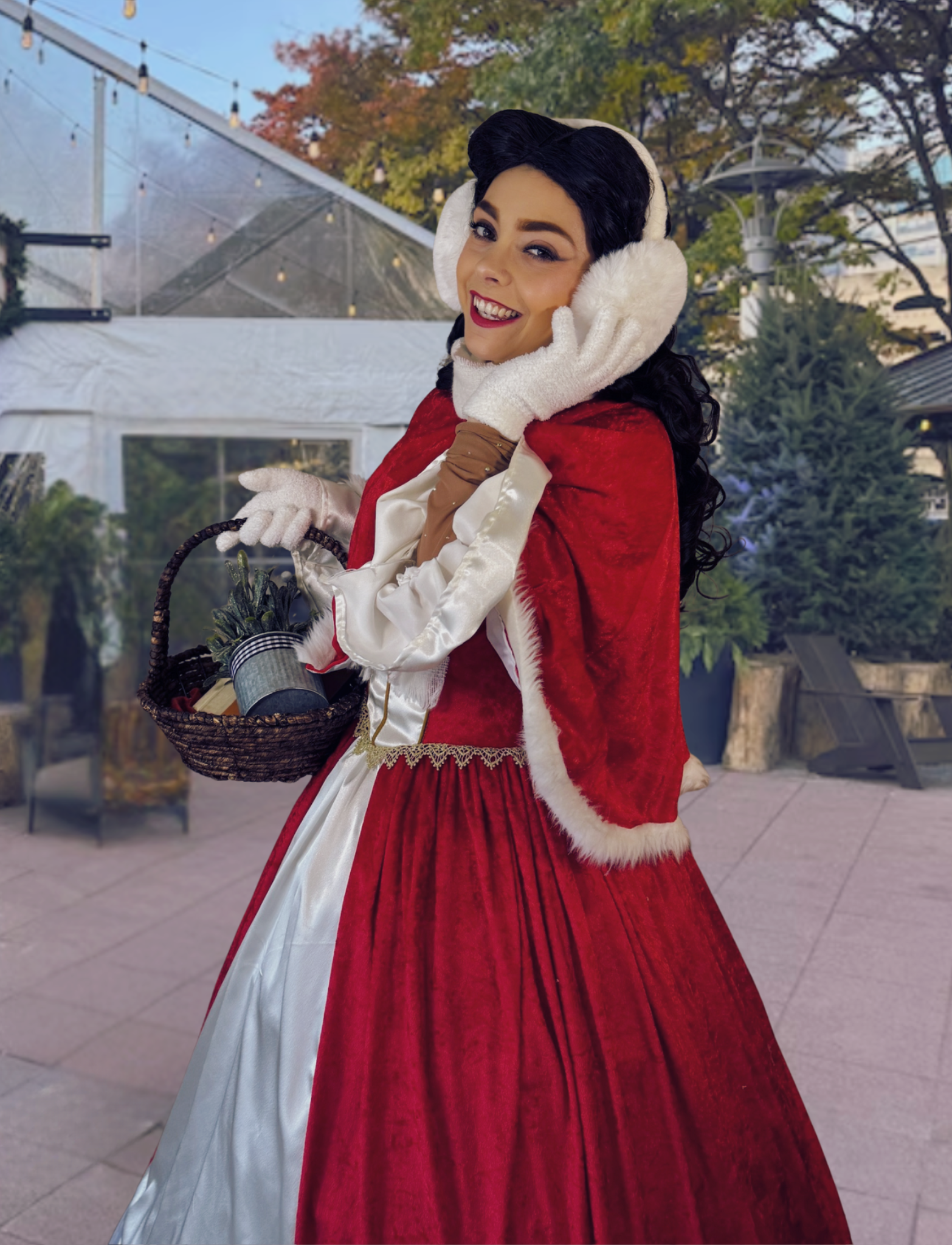 Woman dressed as Mrs. Claus, holding a basket with Christmas decorations, smiling and touching her face with white gloves, outdoors with trees and string lights in the background.