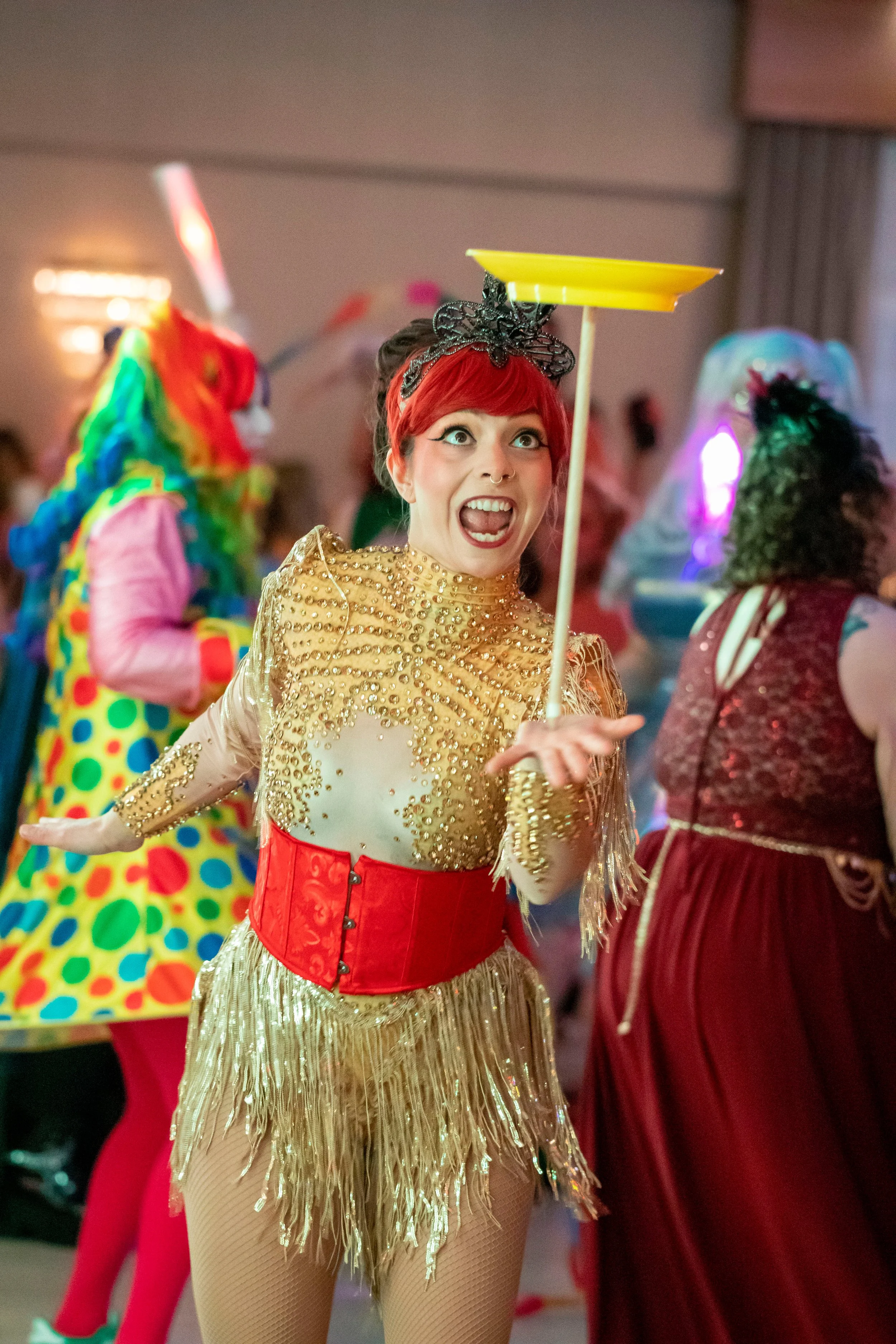 A young woman is balancing a spinning yellow plate on her hand, with bright red hair and a matching red corset