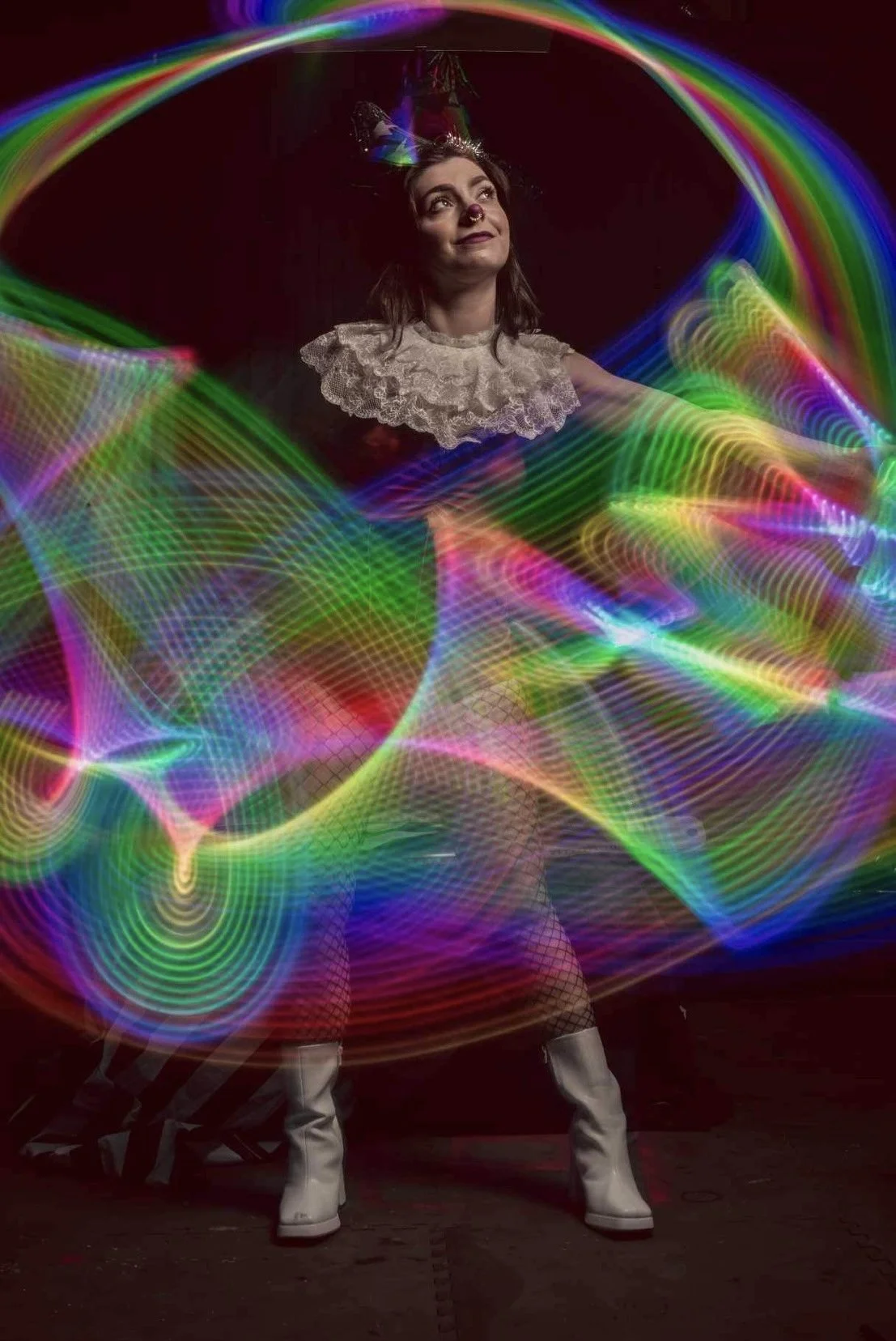 A woman dressed in a whimsical clown costume with a ruffled collar, clown makeup, and white boots, surrounded by colorful light trailscreating a playful and magical scene.