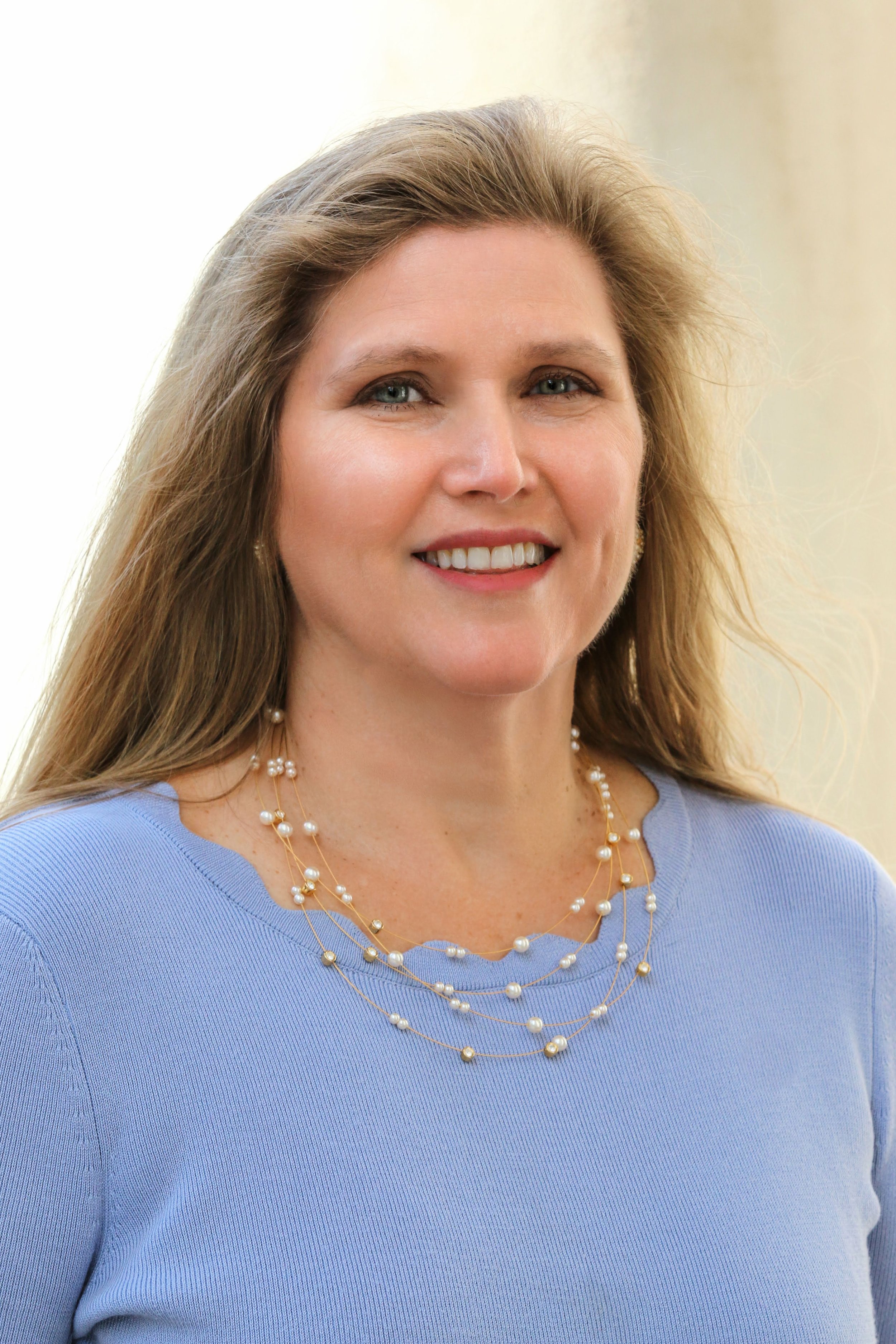 A woman with long blonde hair wearing a light blue top and layered pearl necklaces, smiling.