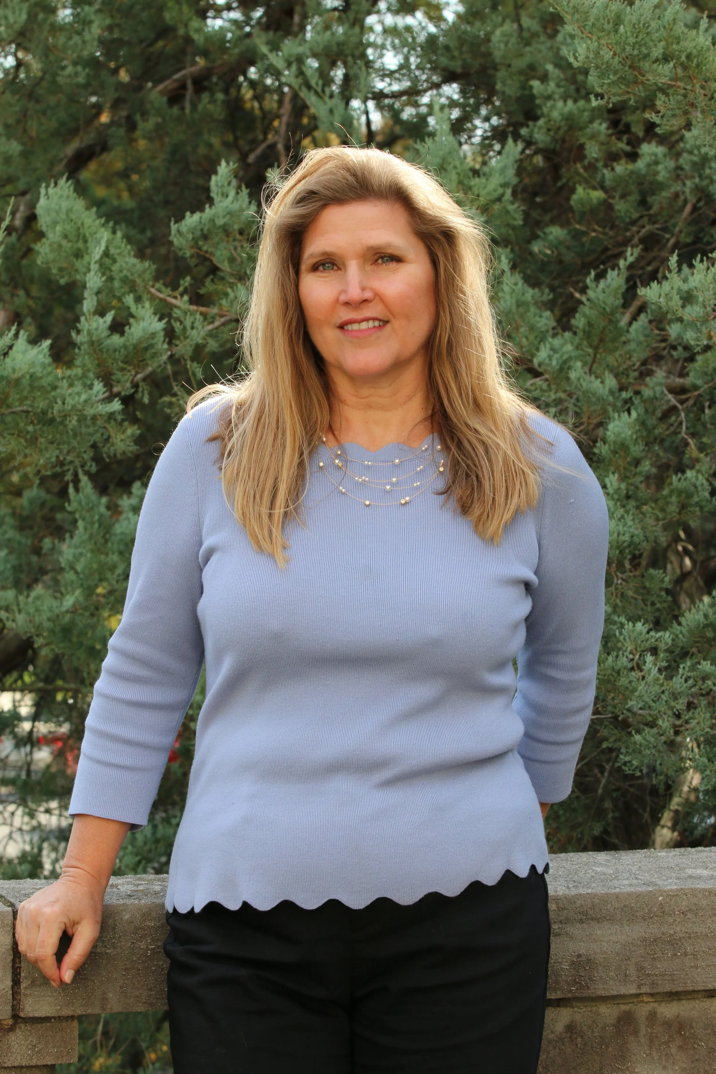 A woman with long blonde hair wearing a light blue sweater and black pants, standing outdoors in front of green trees, leaning on a stone ledge.
