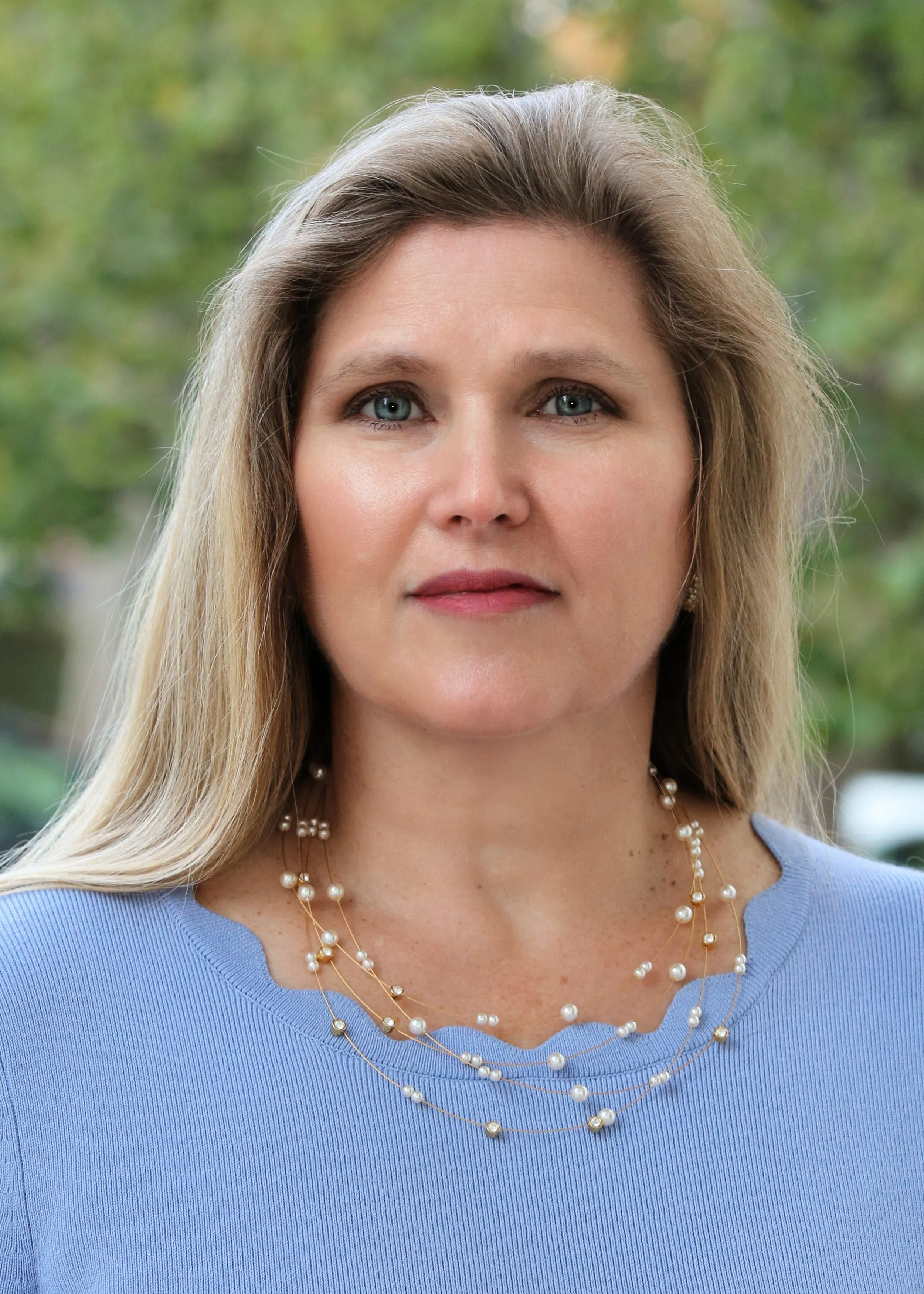 A woman with blonde hair and blue eyes, wearing a light blue top and layered pearl necklaces, stands outdoors with blurred green foliage in the background.