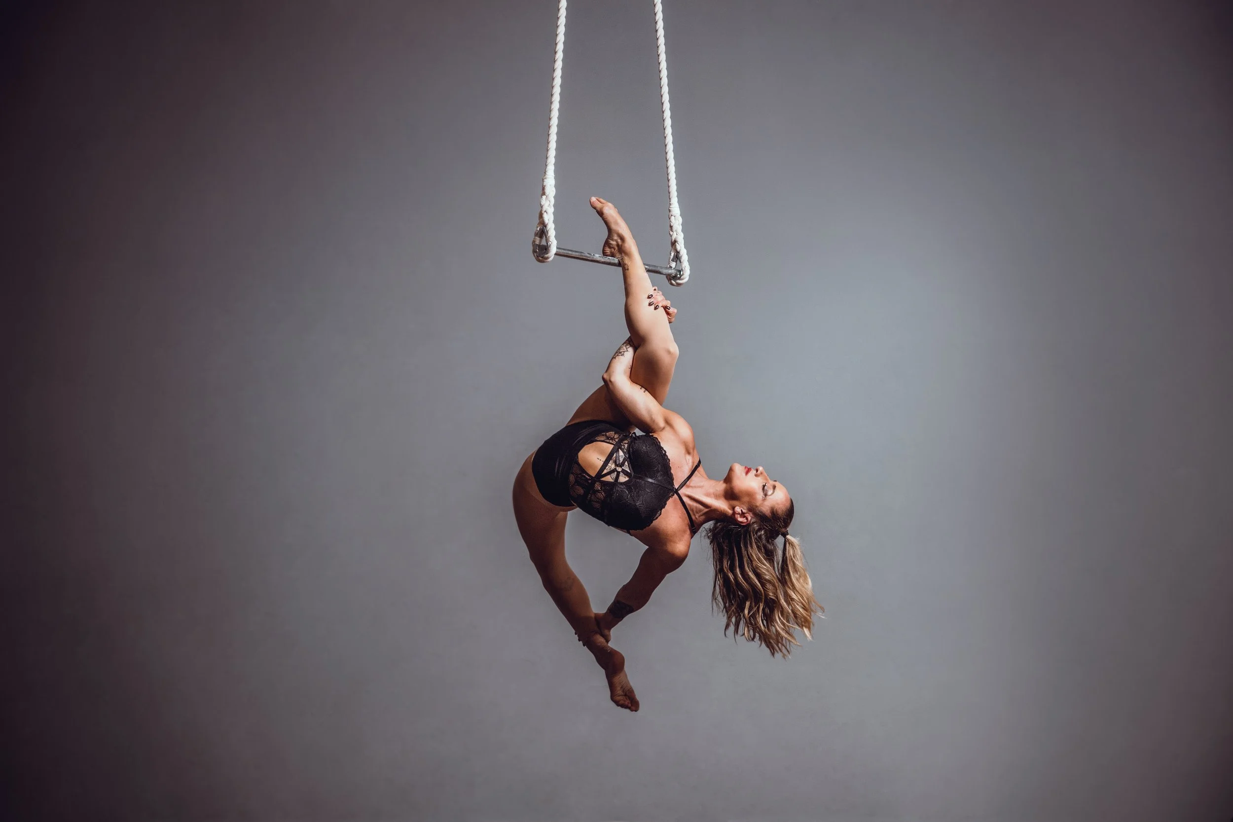 A woman performing aerial silk acrobatics on a trapeze, hanging upside down with her legs bent and her head near the ground, wearing black lingerie against a neutral gray background.