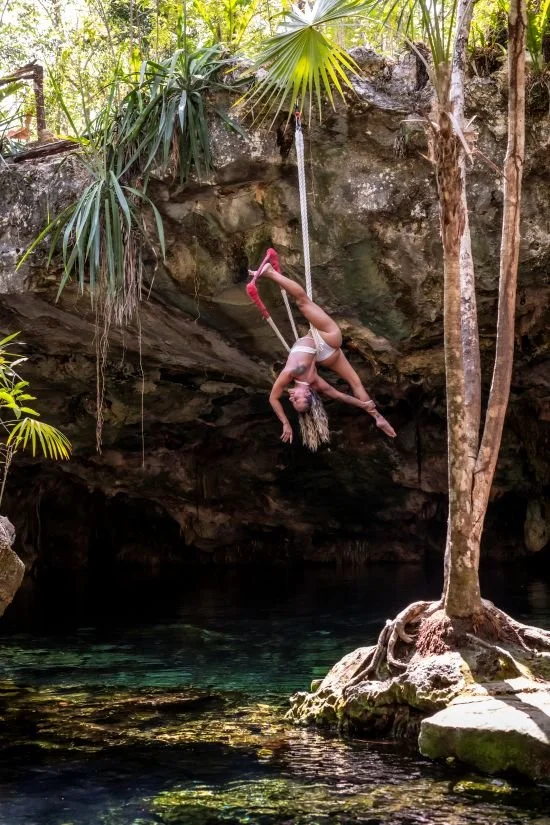 A woman in a white top and shorts hanging upside down from a rope swing over a natural water pool in a lush jungle setting.