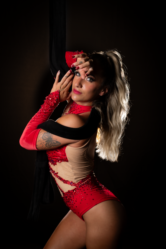A woman with long, wavy blonde hair poses with her right arm raised and her hand near her face against a dark background. She wears a red and beige costume with embellishments, including long red gloves with decorative elements, revealing her tattooed arm. She looks directly at the camera with a confident expression.