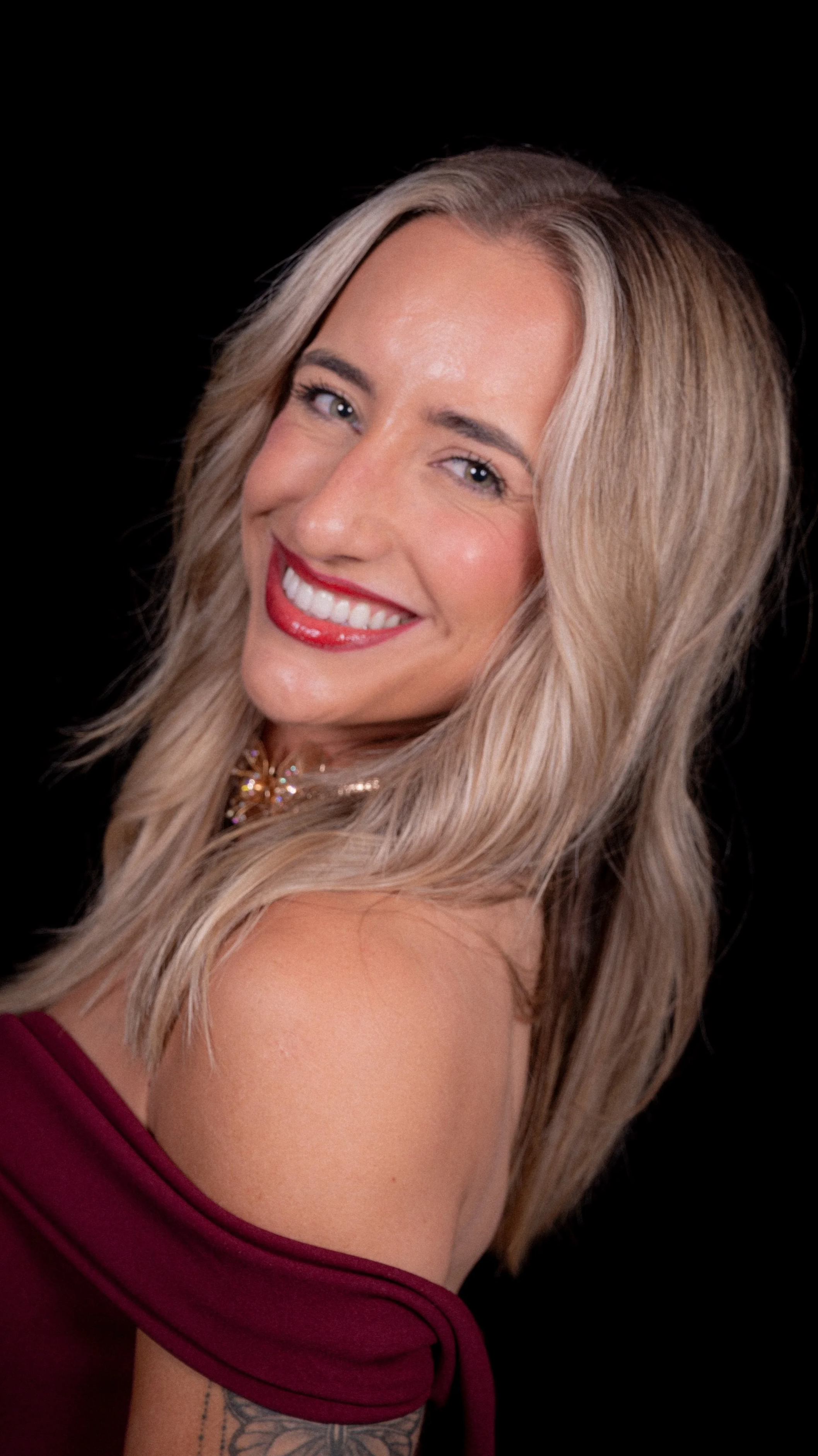 Smiling woman with blonde hair and red lipstick, wearing a burgundy off-shoulder top, against a black background.