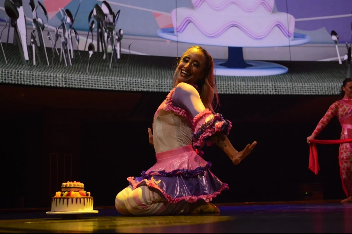 A woman kneeling on stage, smiling, with a birthday cake with fruit slices in front of her, and another performer in the background holding a red cloth.