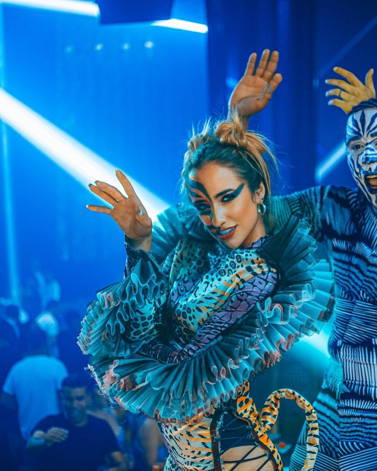 A woman dressed in a colorful animal print costume, with face paint resembling a tiger, posing confidently with a hand near her face in a lively, illuminated setting with blue lighting and a person with similar face paint in the background.