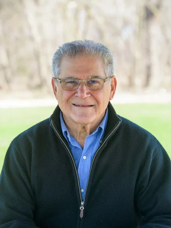 A smiling elderly man with gray hair and glasses, wearing a blue shirt and black zip-up jacket, outdoors in a park-like setting.