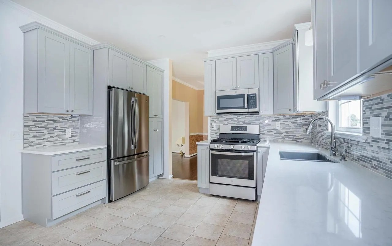 Modern kitchen with white cabinets, stainless steel refrigerator, gas stove, microwave, and a black sink, beige tiled floor, and a gray backsplash.