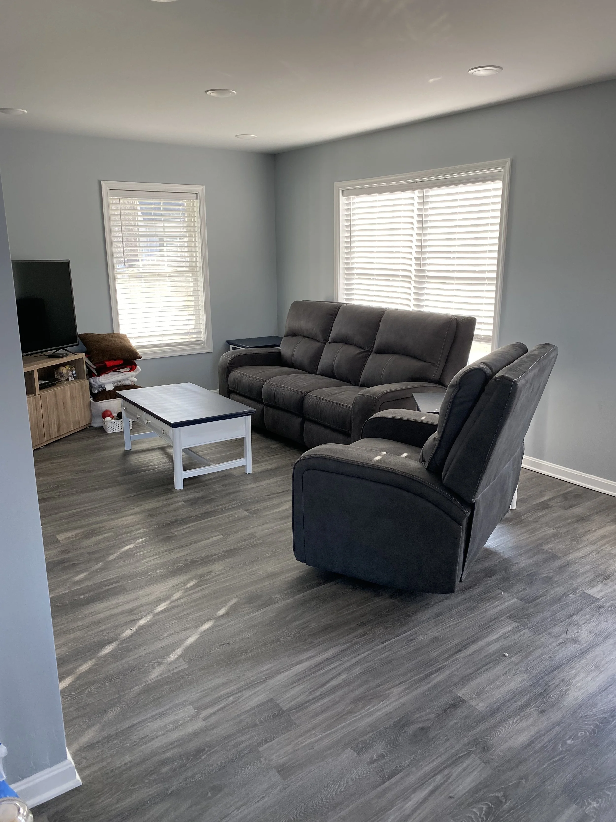 Living room with gray walls, hardwood flooring, a black sofa, a recliner, a small white and blue coffee table, a TV on a wooden stand, and windows with white blinds.