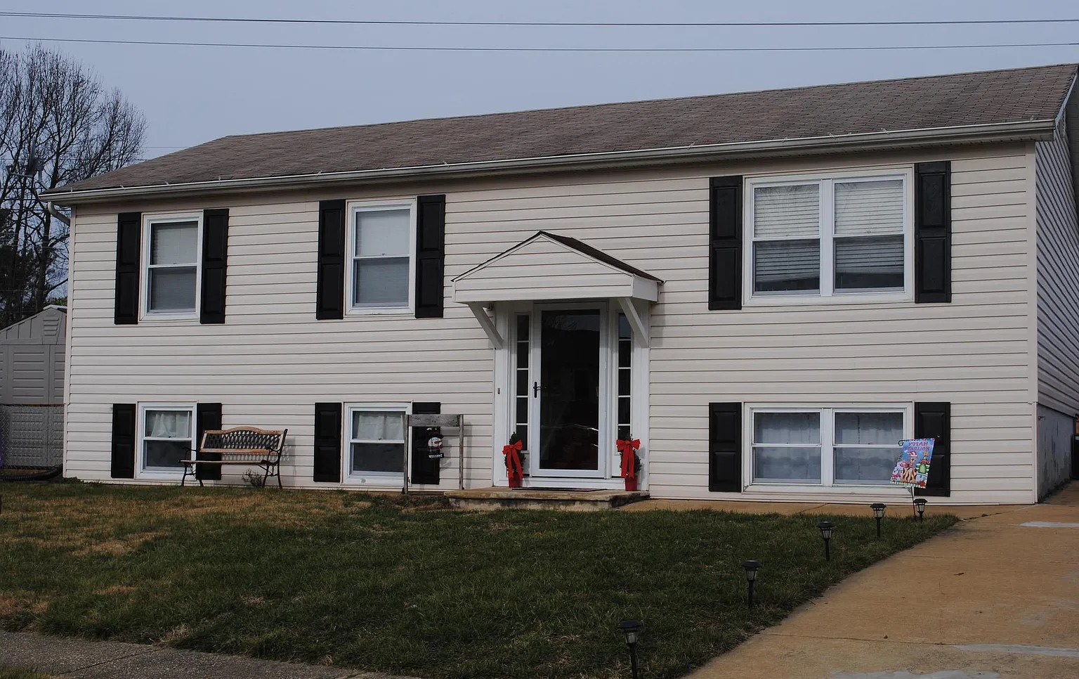 Front view of a two-story house with white siding, black shutters, and a front door decorated for Christmas with red bows. The house has a small front porch, a bench, and a sidewalk leading to it, with a lawn and outdoor lighting along the walkway.