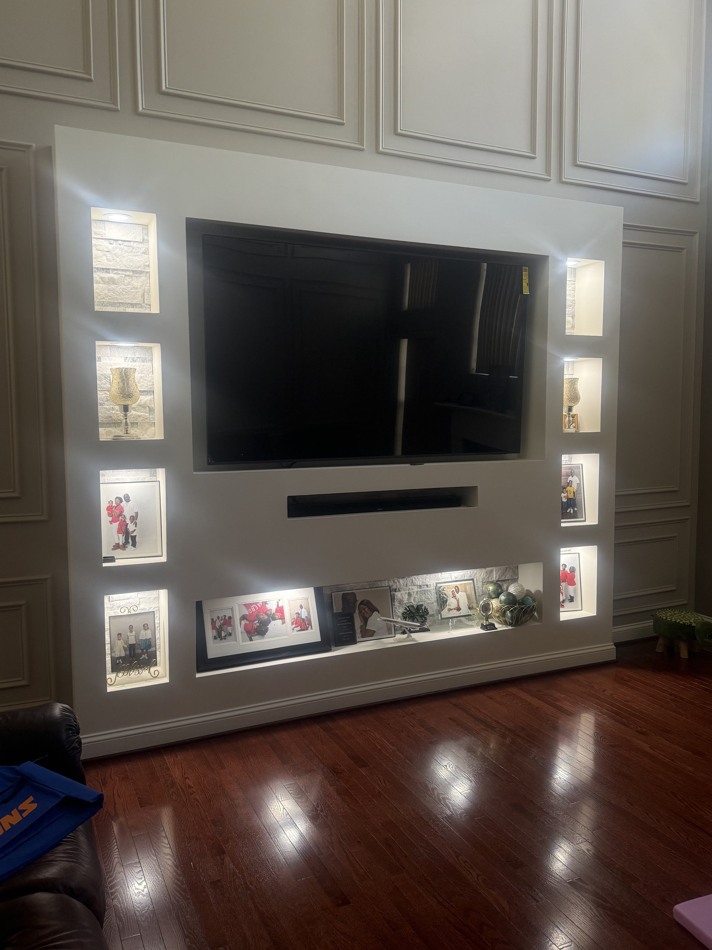 Living room entertainment wall with a large flat-screen TV, illuminated display niches on each side holding small decorative items and photos, and framed photographs below on built-in shelving.