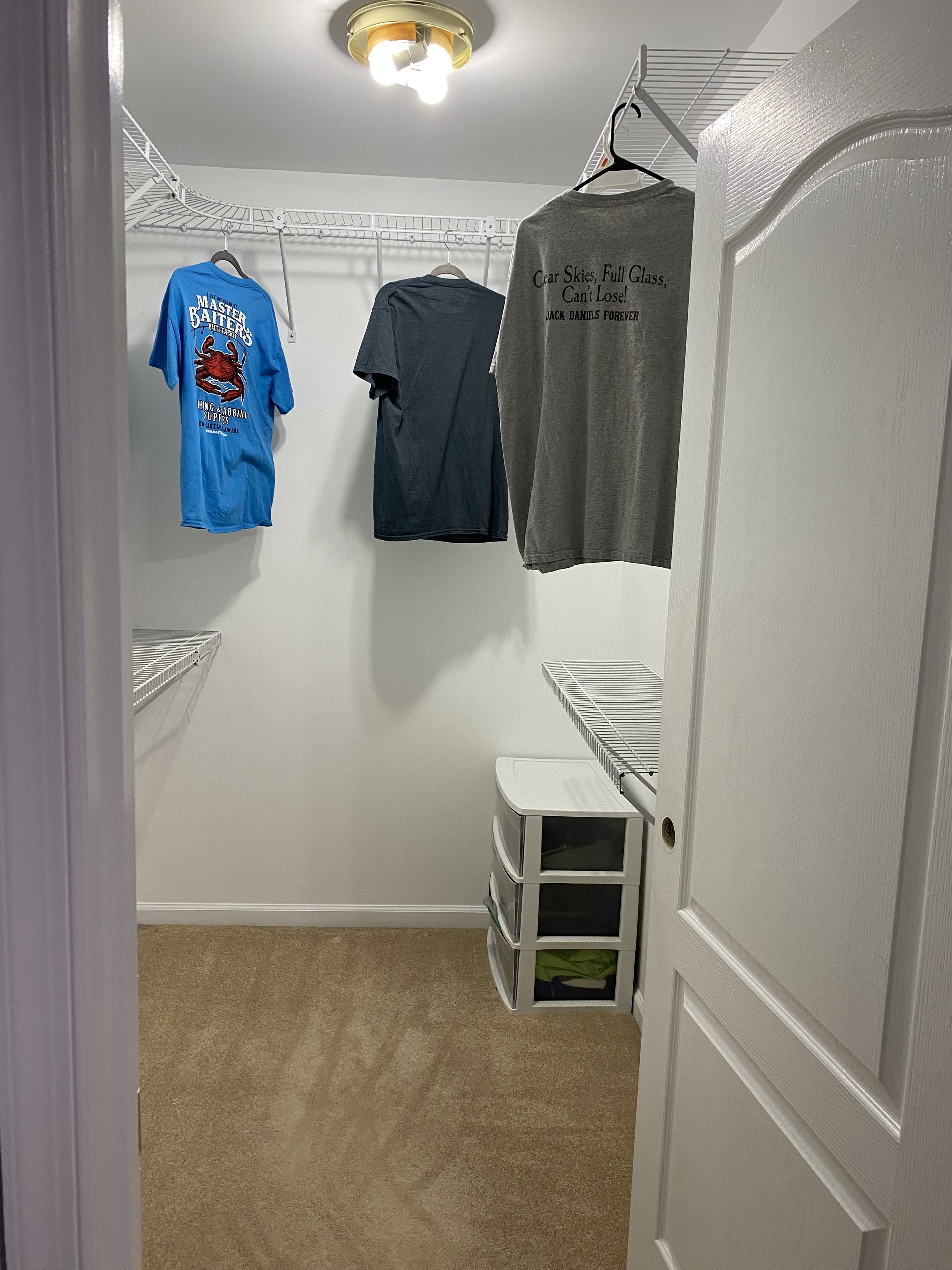 Empty walk-in closet with three shirts hanging from a wire shelf, a small white storage unit with three drawers, and beige carpet flooring.