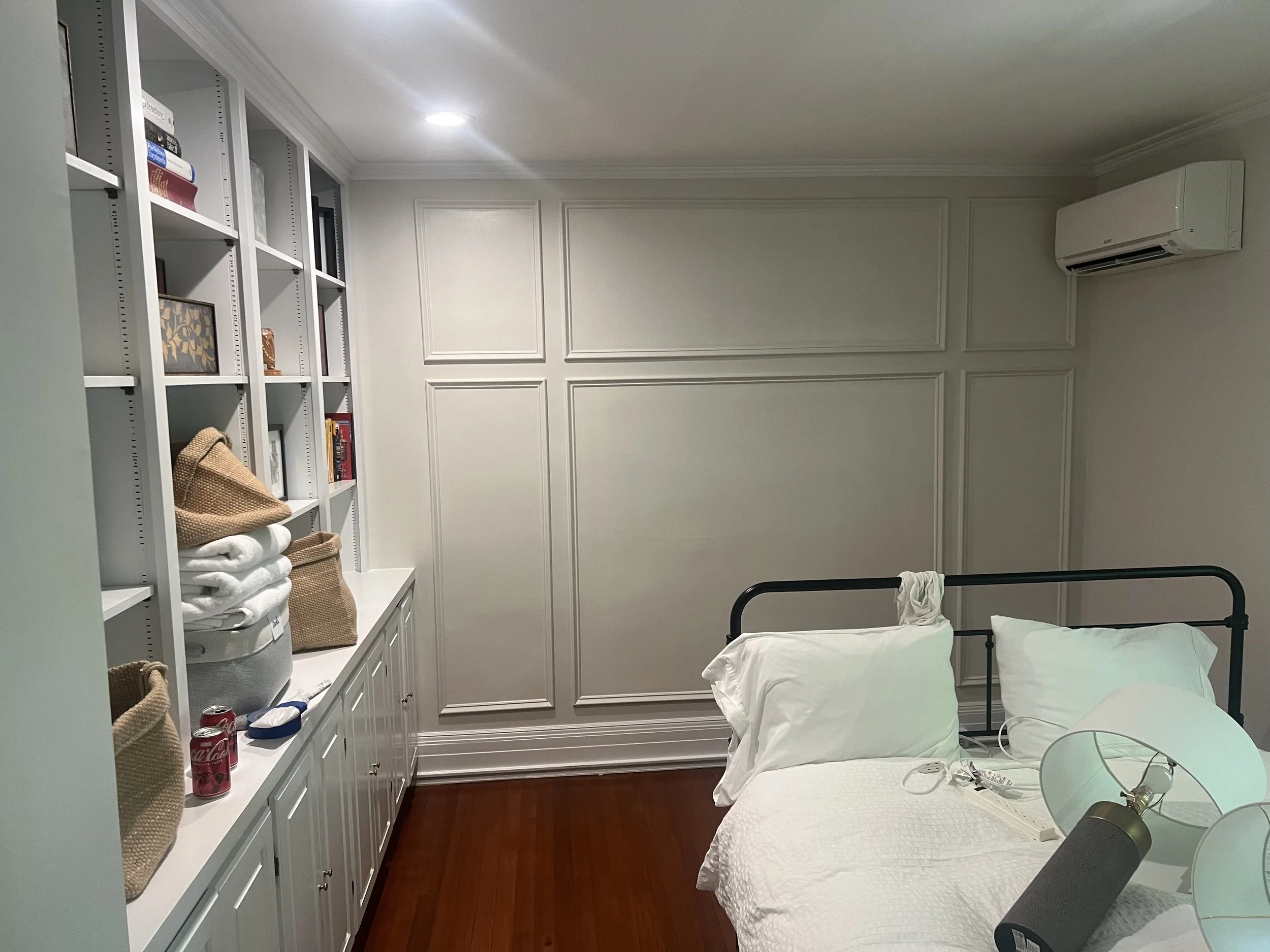 A bedroom with white walls, a white bed with pillows, a lamp, and a wall-mounted air conditioner. There is a white bookshelf on the left side with books, towels, and bags. The room has hardwood floors.