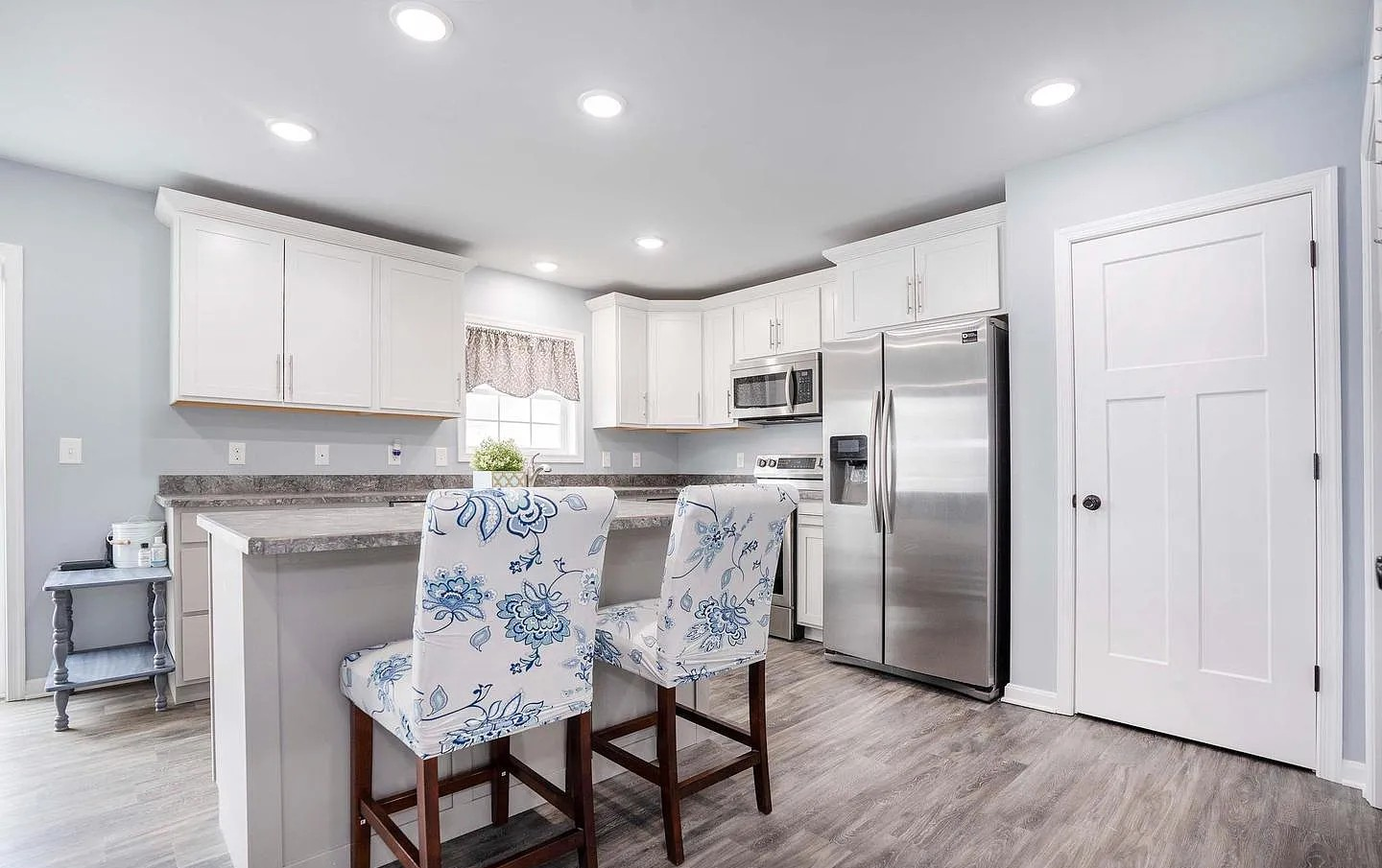 A bright, modern kitchen with white cabinets, stainless steel appliances, a marble countertop island, and two floral-patterned barstools with wooden legs. There's a small window with a valance above the sink and recessed ceiling lights.