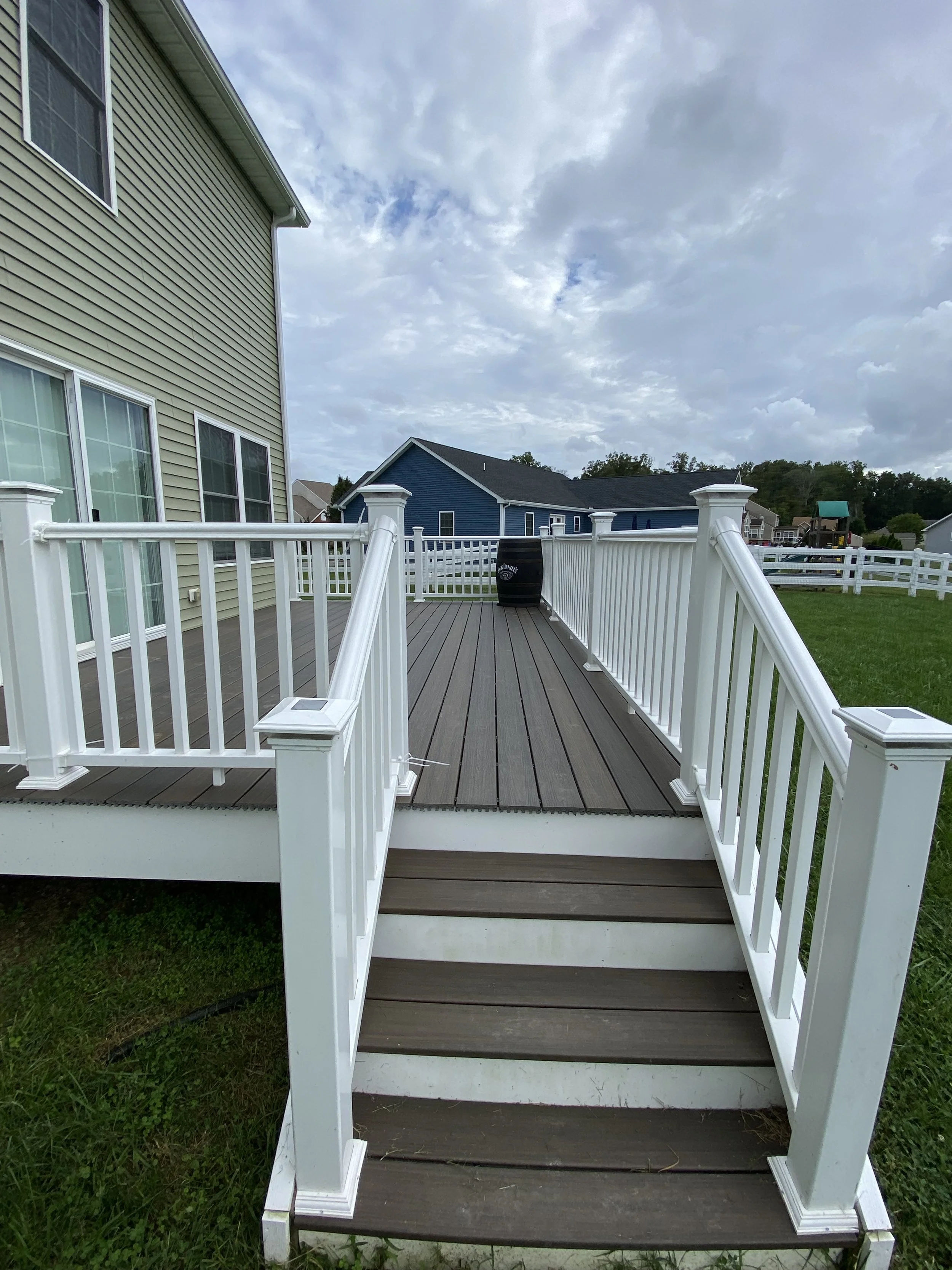 A view of a backyard wooden deck with white railing, accessed by three wooden steps, attached to a yellow house. In the distance, there are other houses, a green lawn, and a cloudy sky.