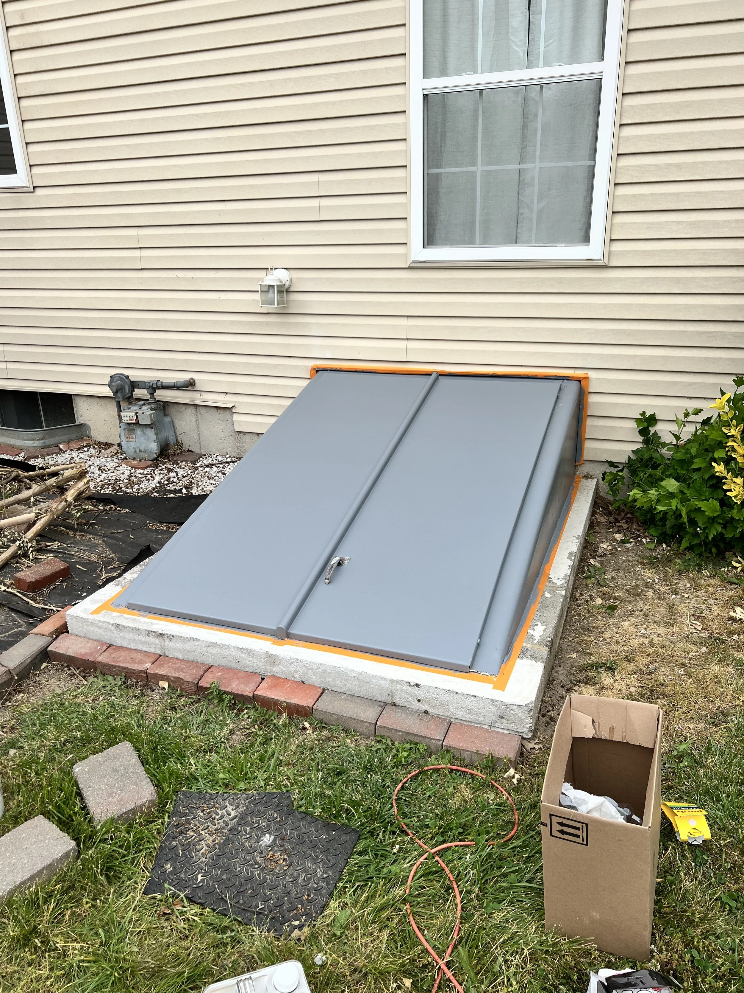 Newly installed gray metal basement hatch door on concrete foundation, surrounded by orange weatherproofing tape, in a yard with grass, bricks, and a cardboard box.