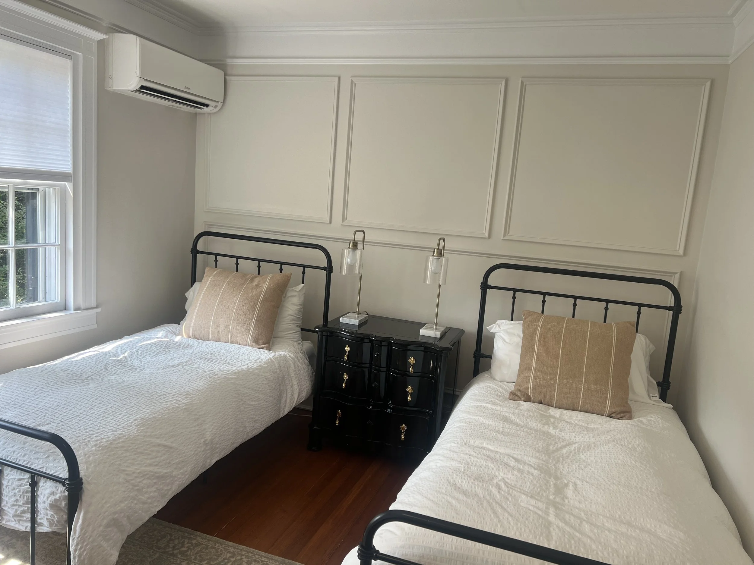 A bedroom with two twin beds with black metal frames, white bedding, and beige pillows. There is a black nightstand between the beds with two glass tabletop lamps. A window with white blinds is on the left, and a white air conditioning unit is mounte