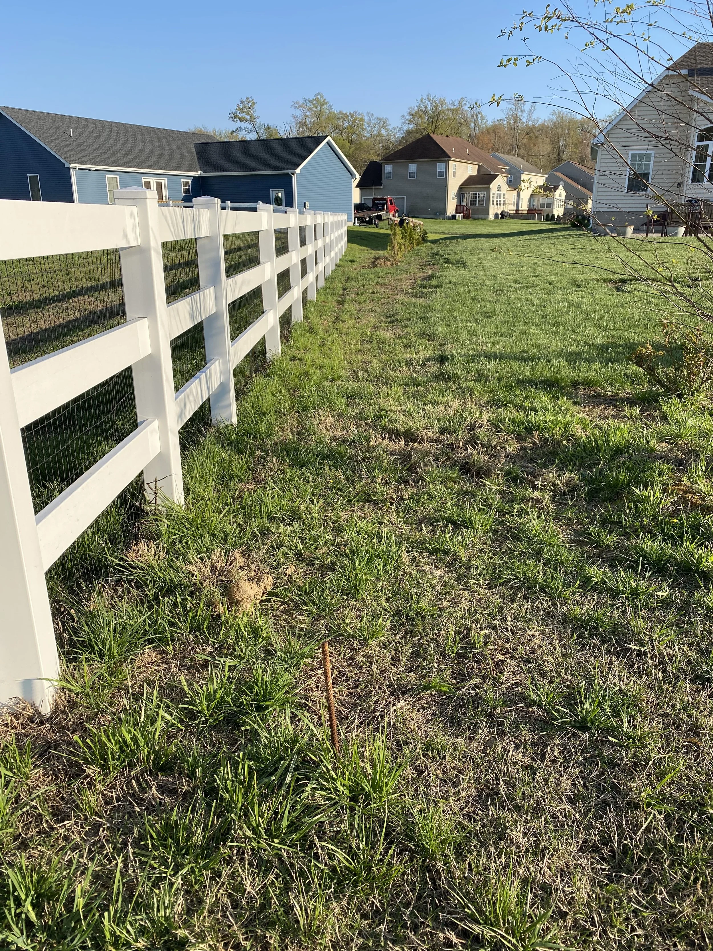 Side yard with green grass, a white fence on the left, and several houses in the background under a clear blue sky.