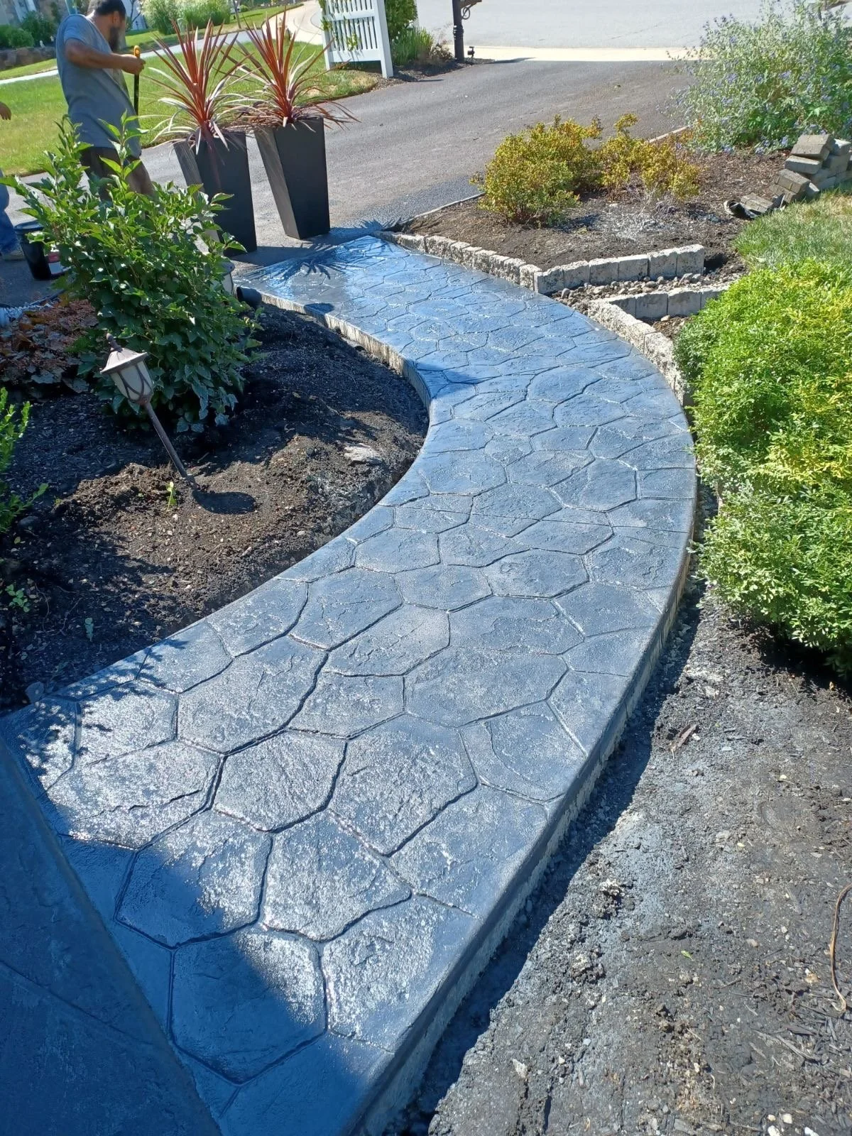 A newly paved arched concrete walkway with a stamped pattern, surrounded by garden beds with plants and shrubs, in front of a driveway and open yard.