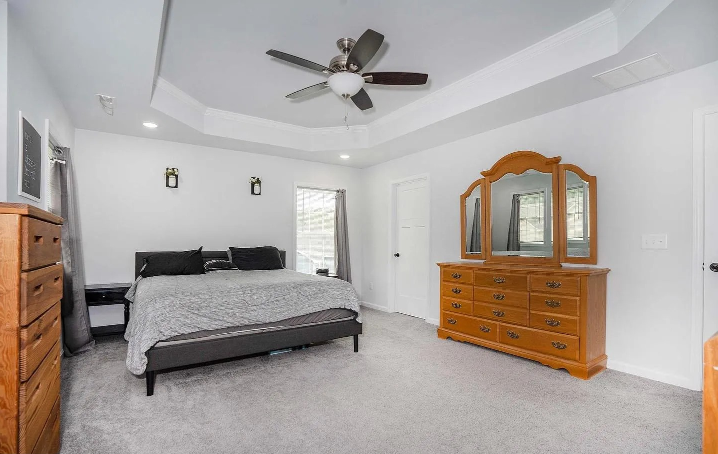 A bedroom with white walls, a ceiling fan, a bed with black pillows, two windows with gray curtains, a wooden dresser with a mirror, and a gray carpet.