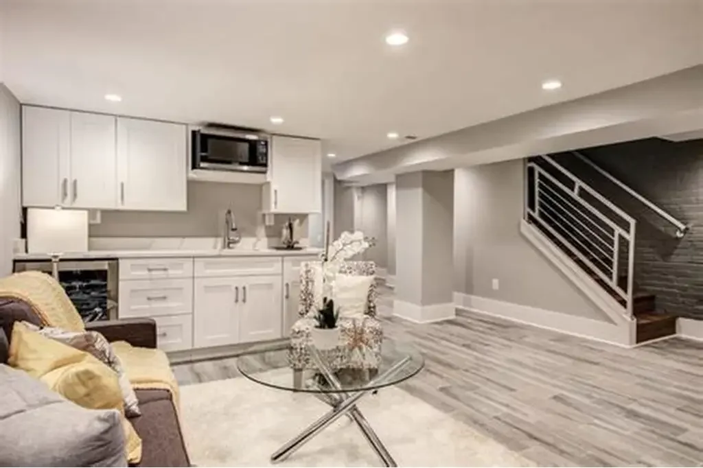 A modern basement with a small kitchenette, a staircase leading upstairs, and a seating area with sofa and decorative pillows.