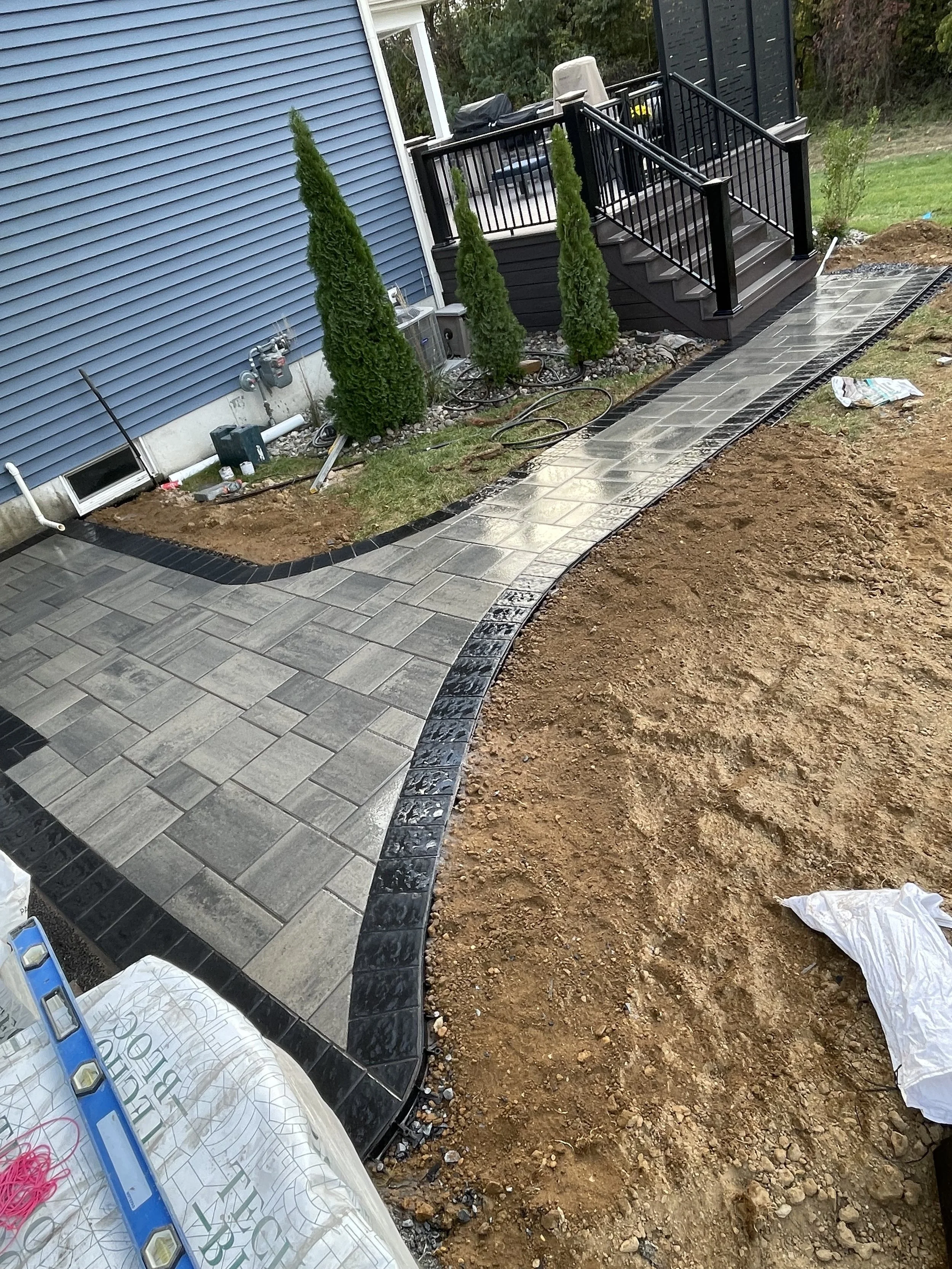 A freshly laid outdoor tile walkway with black border edging, adjacent to a house with blue siding, new green shrubs, and a small wooden staircase with black railing leading to a deck.