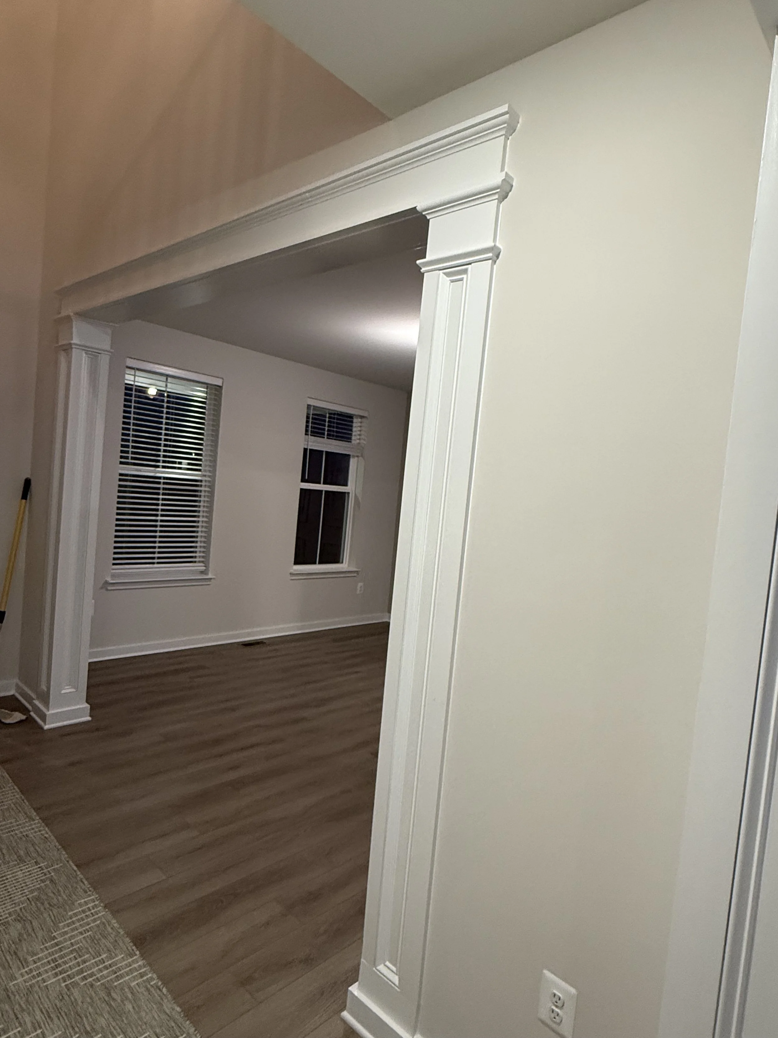 Interior view of a home featuring a white archway with decorative molding, wooden flooring, and windows with blinds.