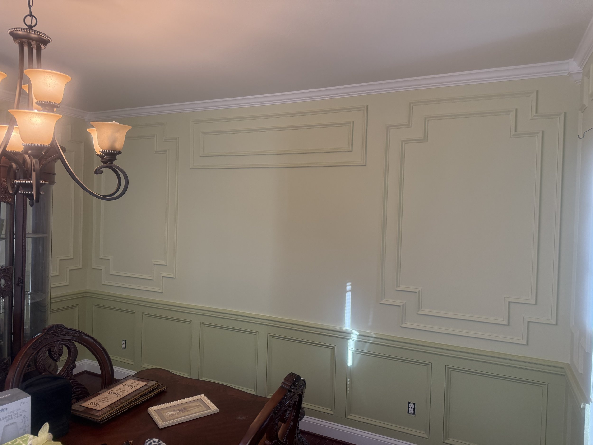 A dining room wall with decorative molding painted in pale green and a chandelier with multiple upward-facing light fixtures. Part of a wooden dining table with plates and other items visible in the lower part of the image.