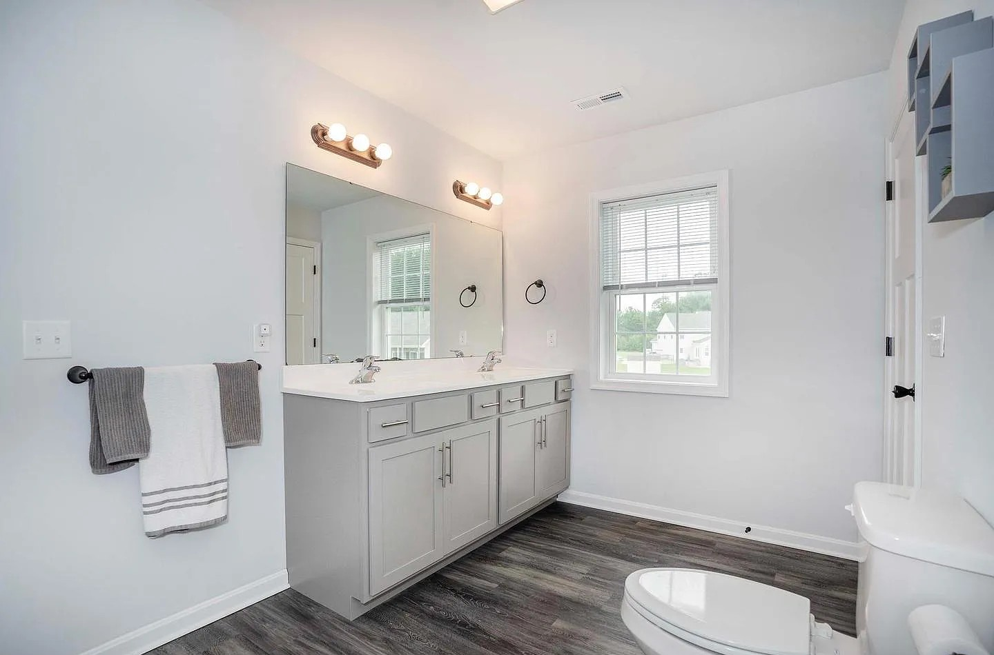 Modern bathroom with gray vanity, double sinks, large mirror, white walls, dark wood flooring, window with blinds, toilet, and wall-mounted lights.