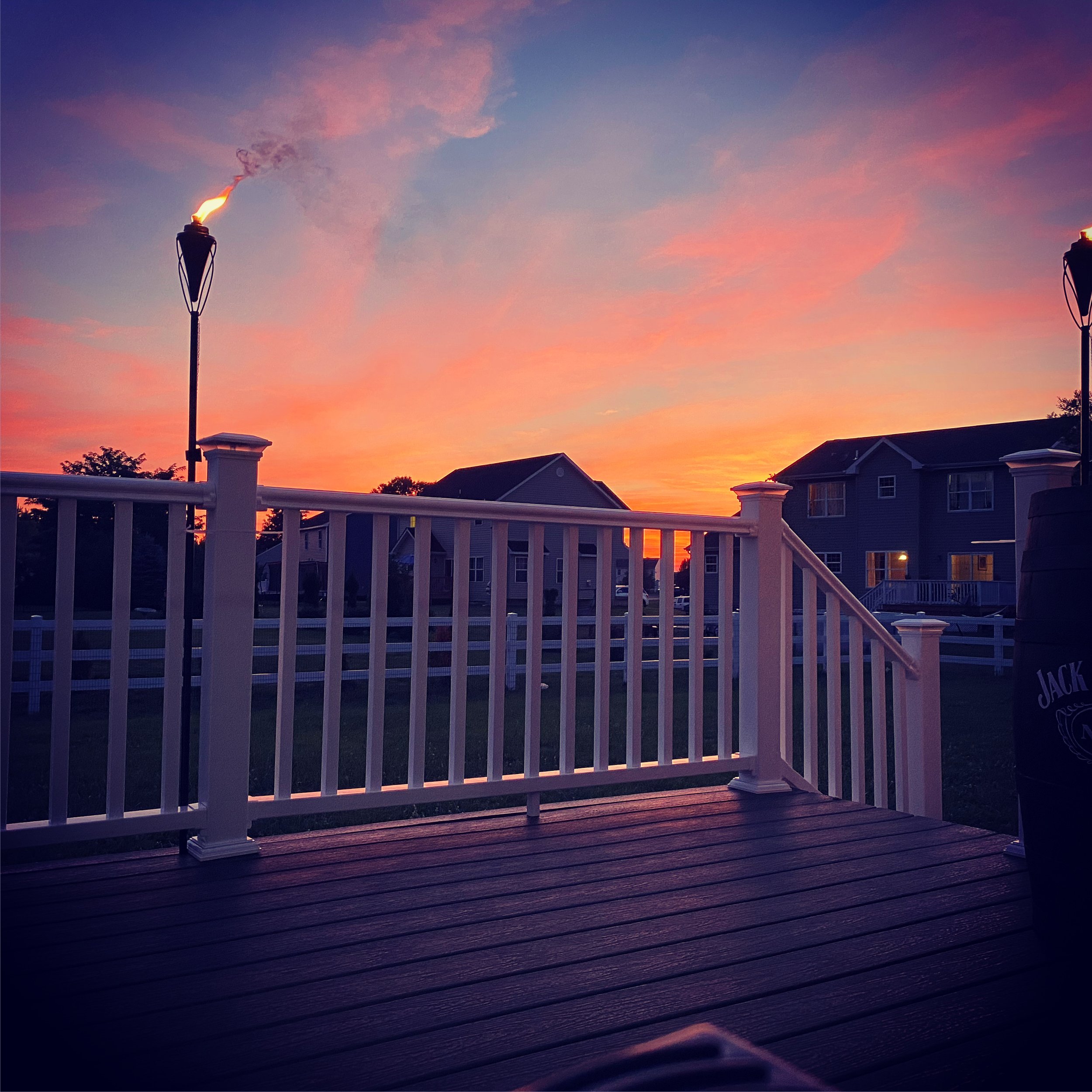 A wooden deck with white railing overlooking a suburban neighborhood at sunset, with a colorful sky and two lampposts emitting light.