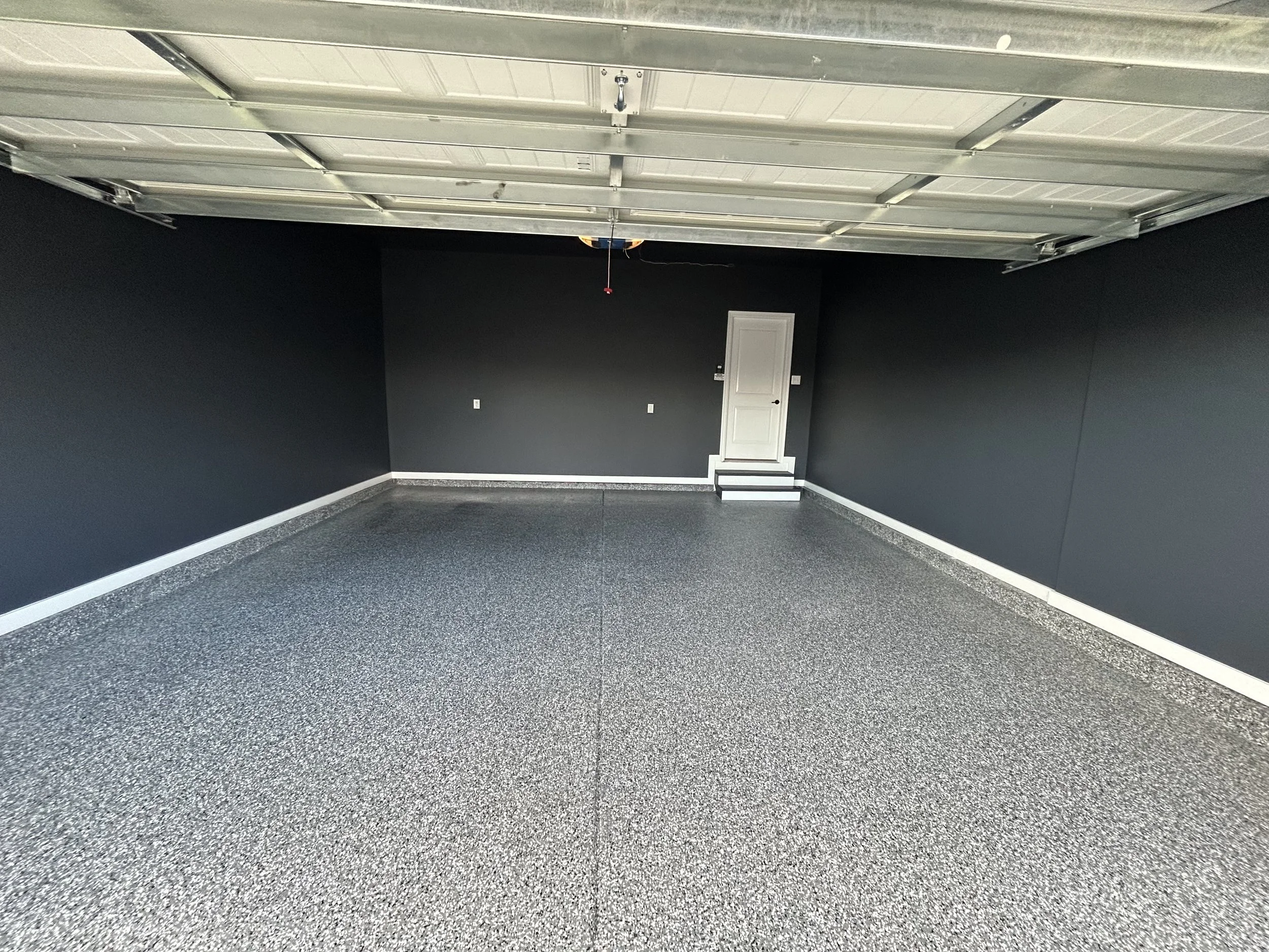 Empty garage with black walls, speckled gray epoxy flooring, and a white door with steps leading up to it.