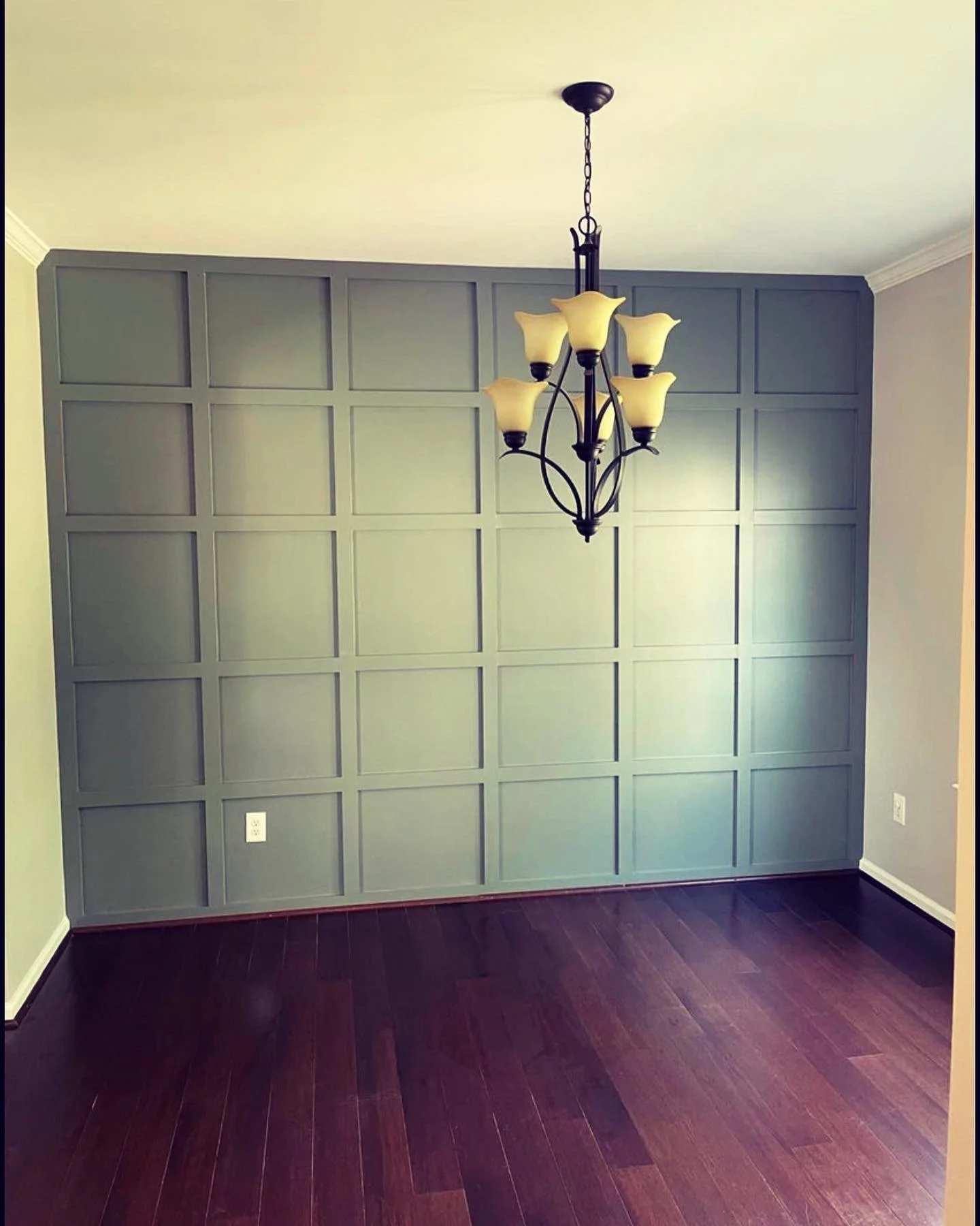 Empty room with a gray paneled accent wall, dark hardwood floor, and a black chandelier with beige glass shades hanging from the ceiling.