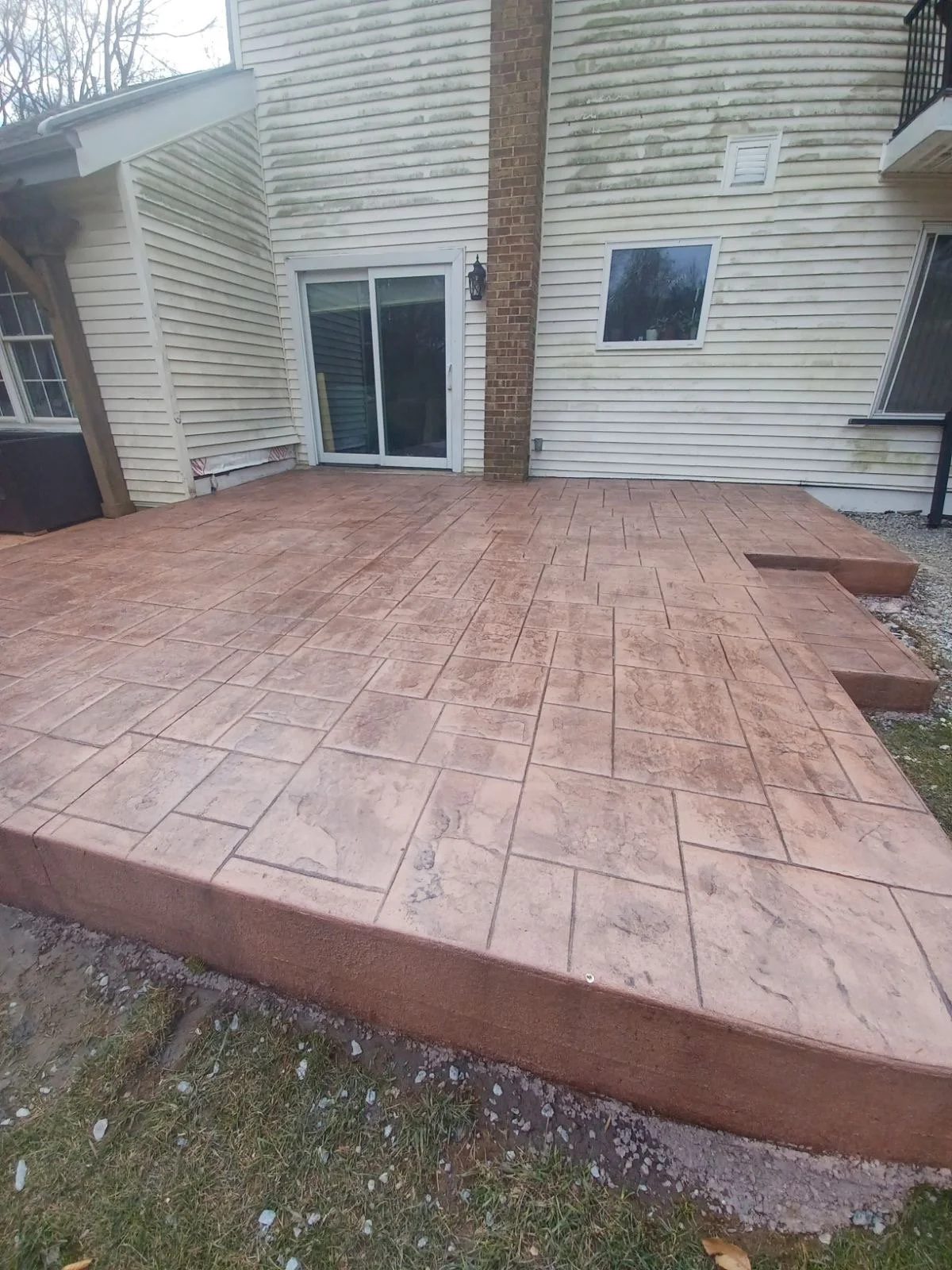 Photo of a freshly paved concrete patio with stamped pattern, attached to a house with siding and a sliding glass door, featuring steps at one end.
