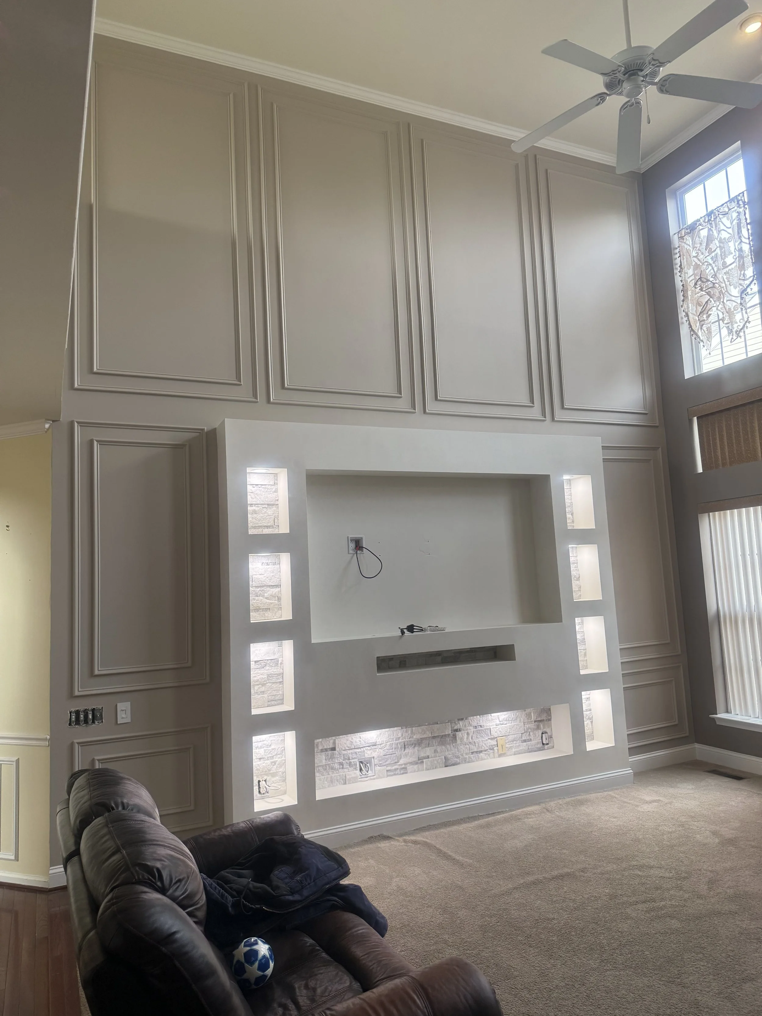 Living room with a large decorative wall featuring built-in shelves and a recessed space for a television, with a ceiling fan, beige carpet, brown leather couch, and large windows with blinds and curtains.
