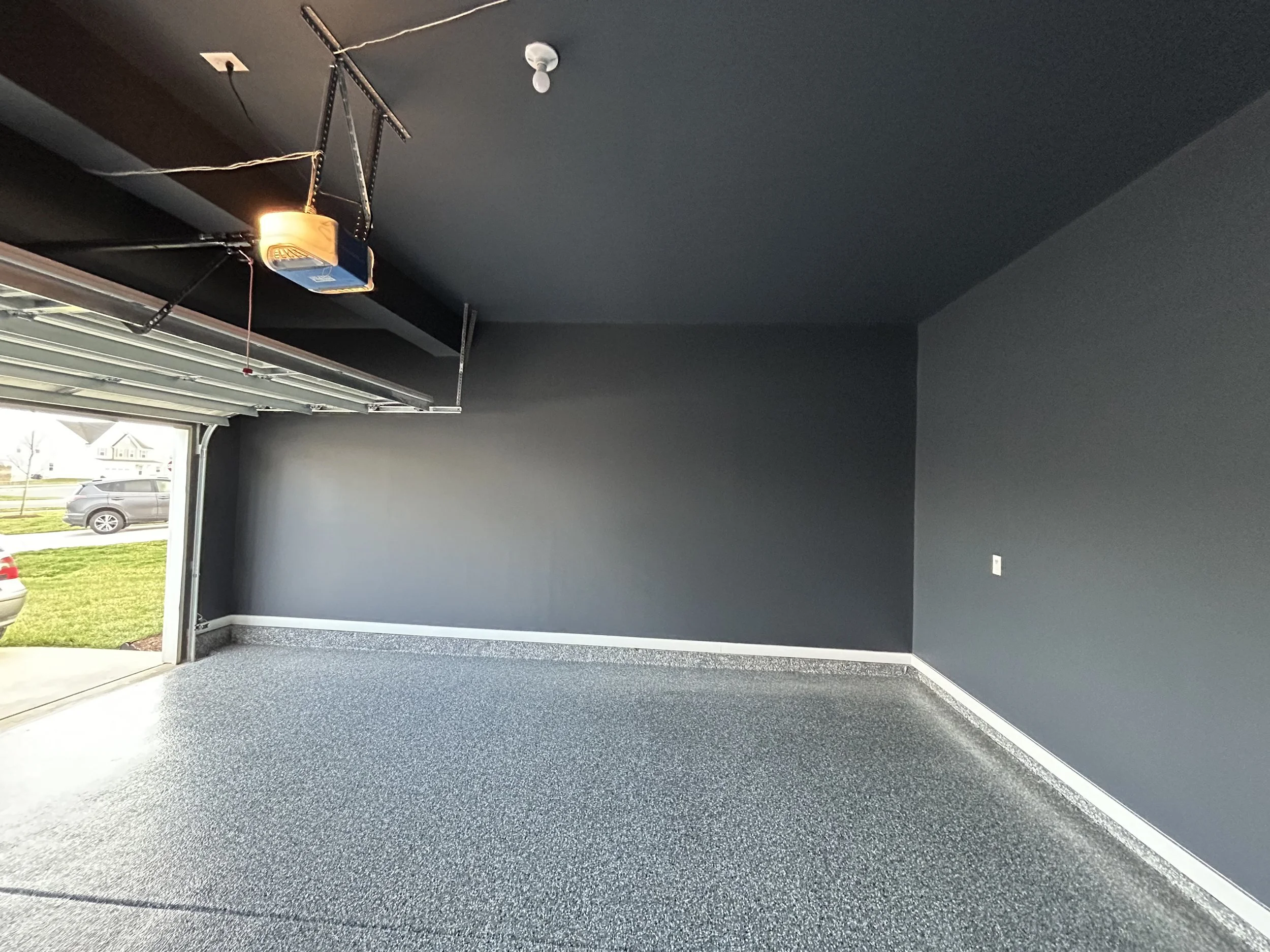 Empty garage with dark painted walls, speckled gray epoxy floor, and an open garage door revealing a residential street with parked cars and houses outside.