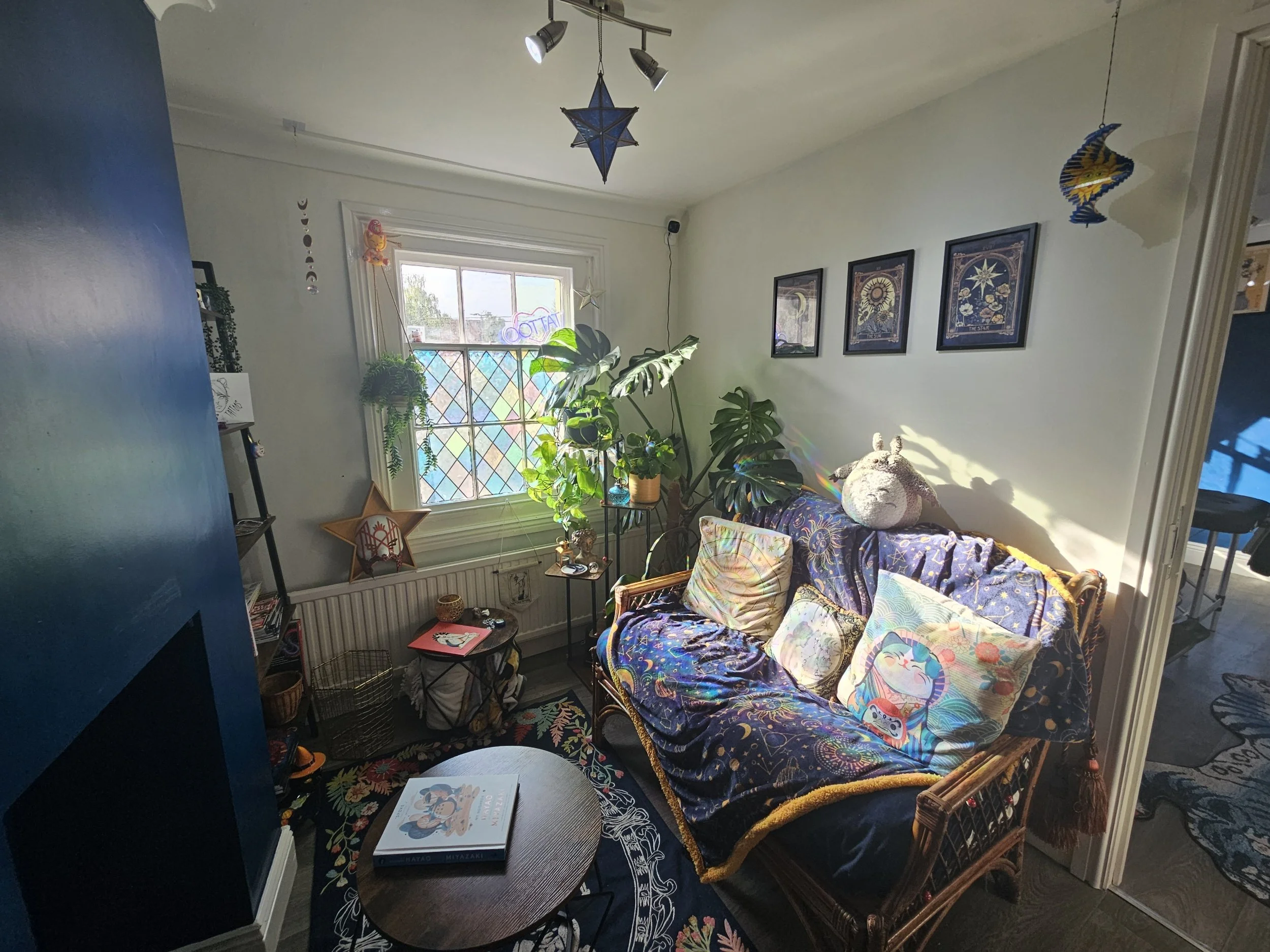 Sunlit living room corner with a wicker sofa covered in a dark blue blanket and colorful pillows, a small round coffee table with a book on top, a window with stained glass and plants, wall art, and decorations hanging from the ceiling.