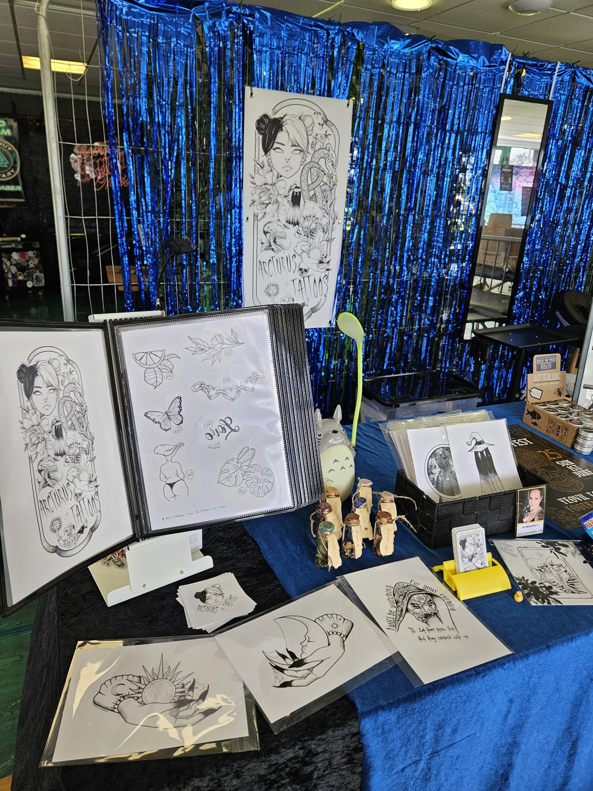Tattoo booth display with tattoo designs, including a drawing of a woman with two buns in her hair, surrounded by flowers, animals, and other images. The booth is decorated with blue metallic streamers and includes tattoo flash sheets, stickers, and small items for sale.