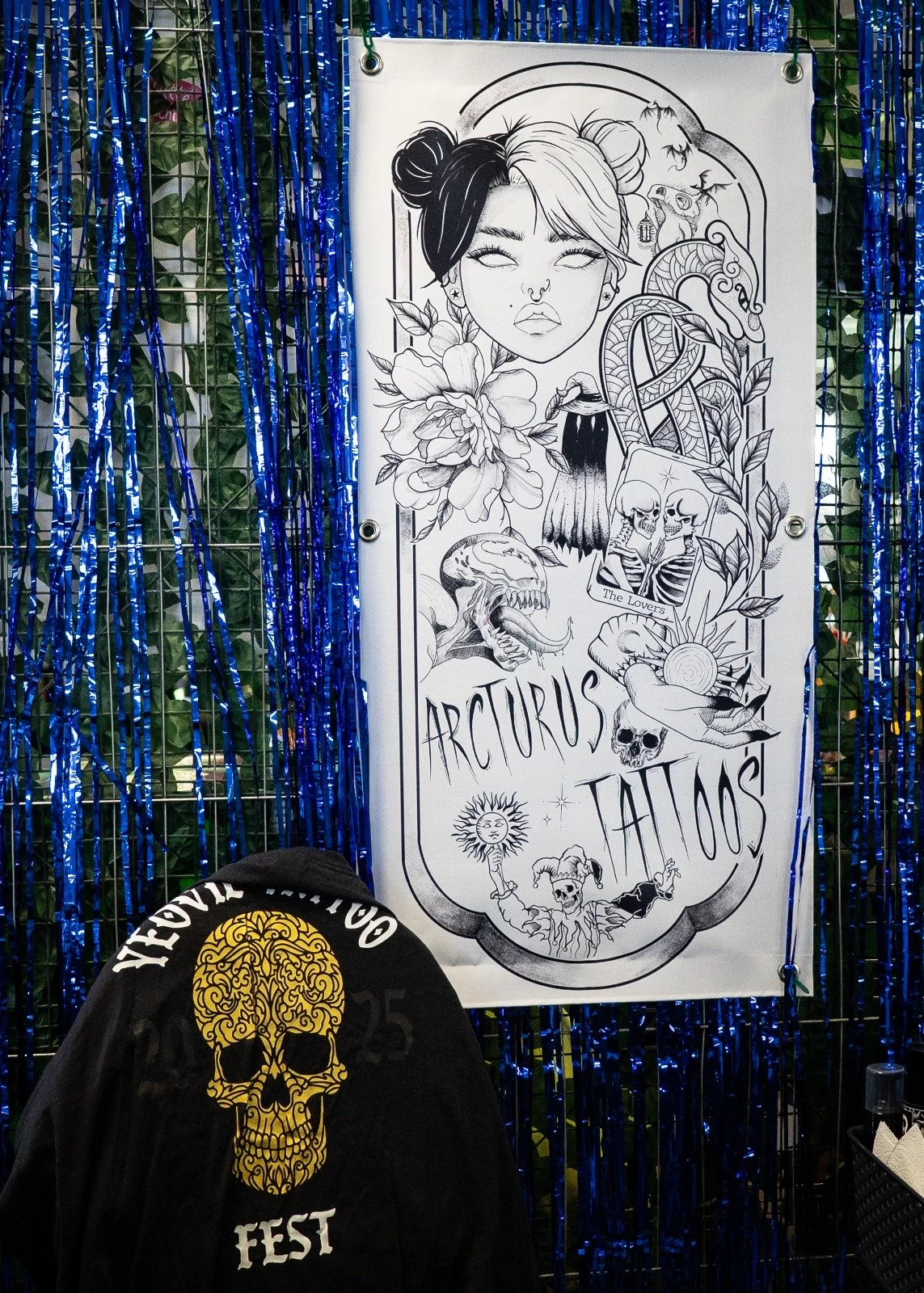 Black hoodie with a yellow skull and floral design in the center, with 'REVEL FEST' written above. Behind it, a large poster displays a line art illustration of a woman's face with floral and fantasy elements, surrounded by zodiac and tarot-themed drawings, and the words 'ARCTURUS TATTOOS'.