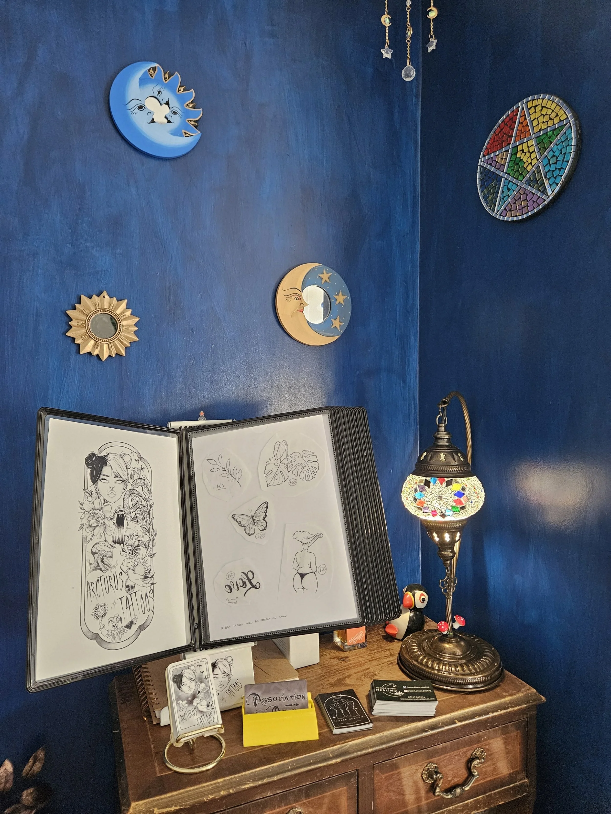 A wooden table with tattoo design sketches, business cards, and small items, against a deep blue wall decorated with moon and sun themed wall art, and a Tiffany-style stained glass lamp.