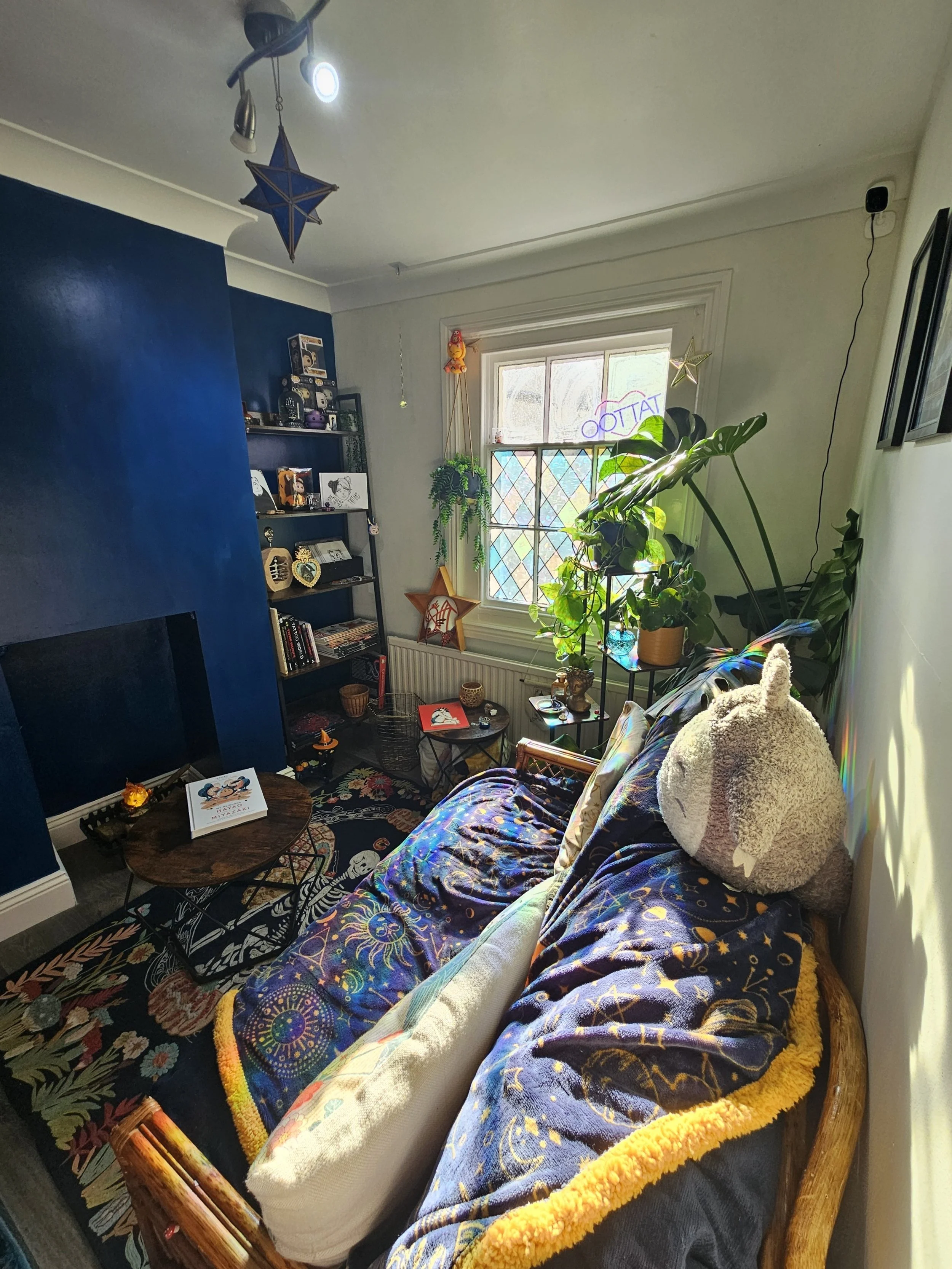 Cozy living room with a dark blue accent wall, a fireplace, a patterned rug, a couch covered with celestial-themed blankets, plush toys, indoor plants on the windowsill, and decorative star ornaments hanging from the ceiling.
