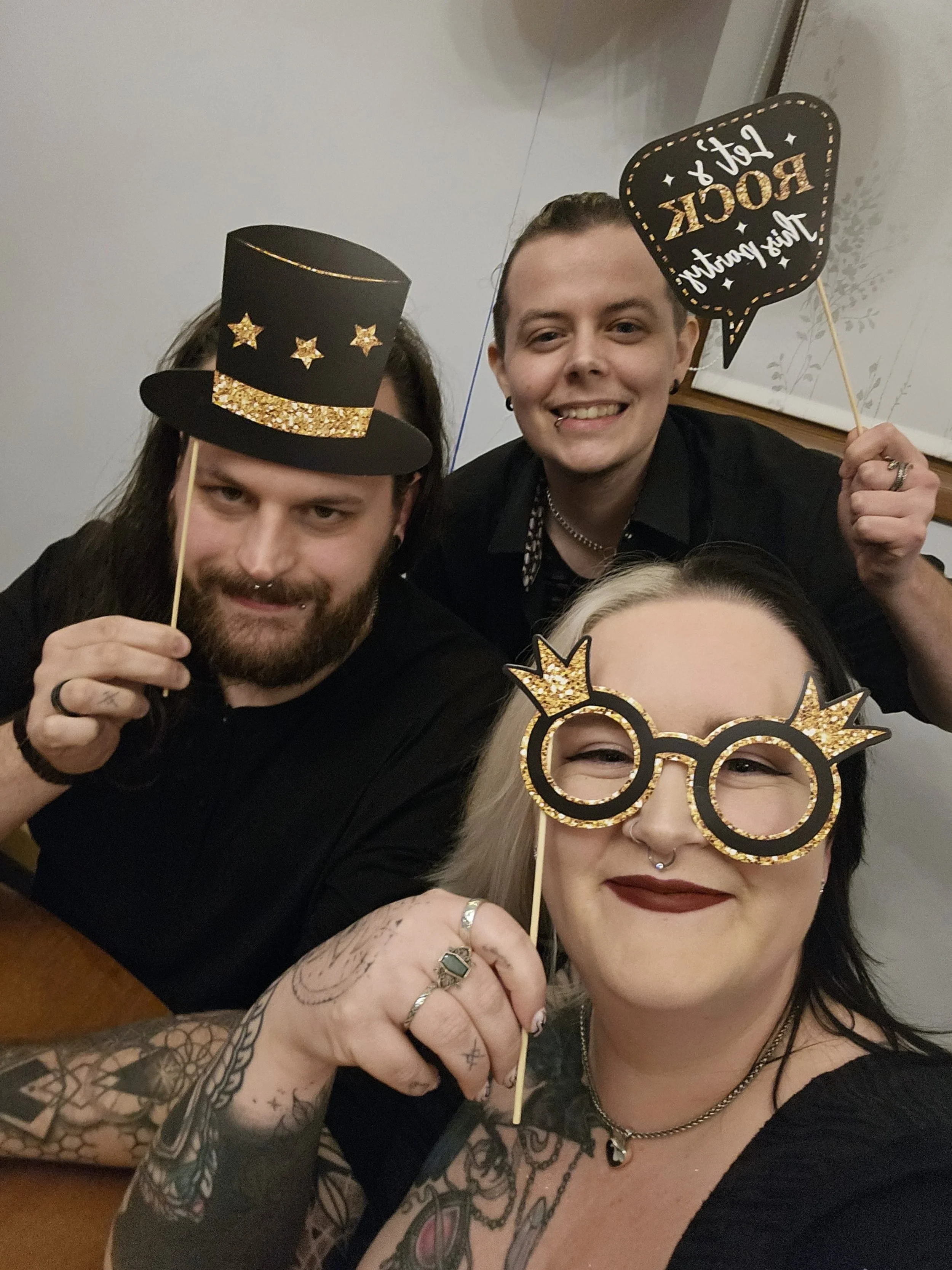 Three people celebrating New Year's Eve, holding festive signs and wearing glittery glasses and hats.