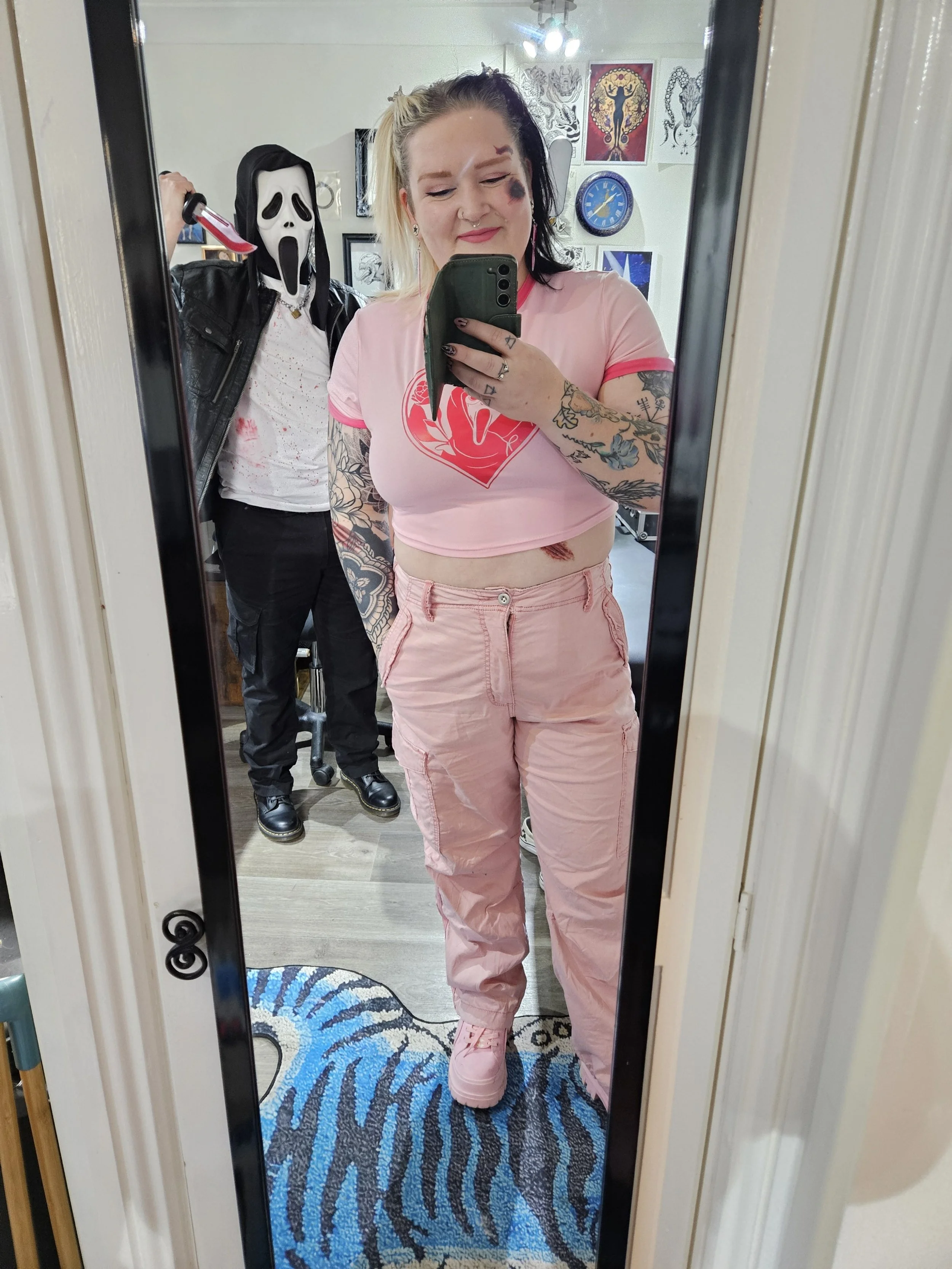 A woman with tattoos taking a mirror selfie in a pink outfit with a pink shirt, pink pants, and pink sneakers, smiling with bleach and black hair. Behind her, a person in a Scream mask, black leather jacket, and holding a fake bloody knife stands in a room decorated with various artwork and posters.