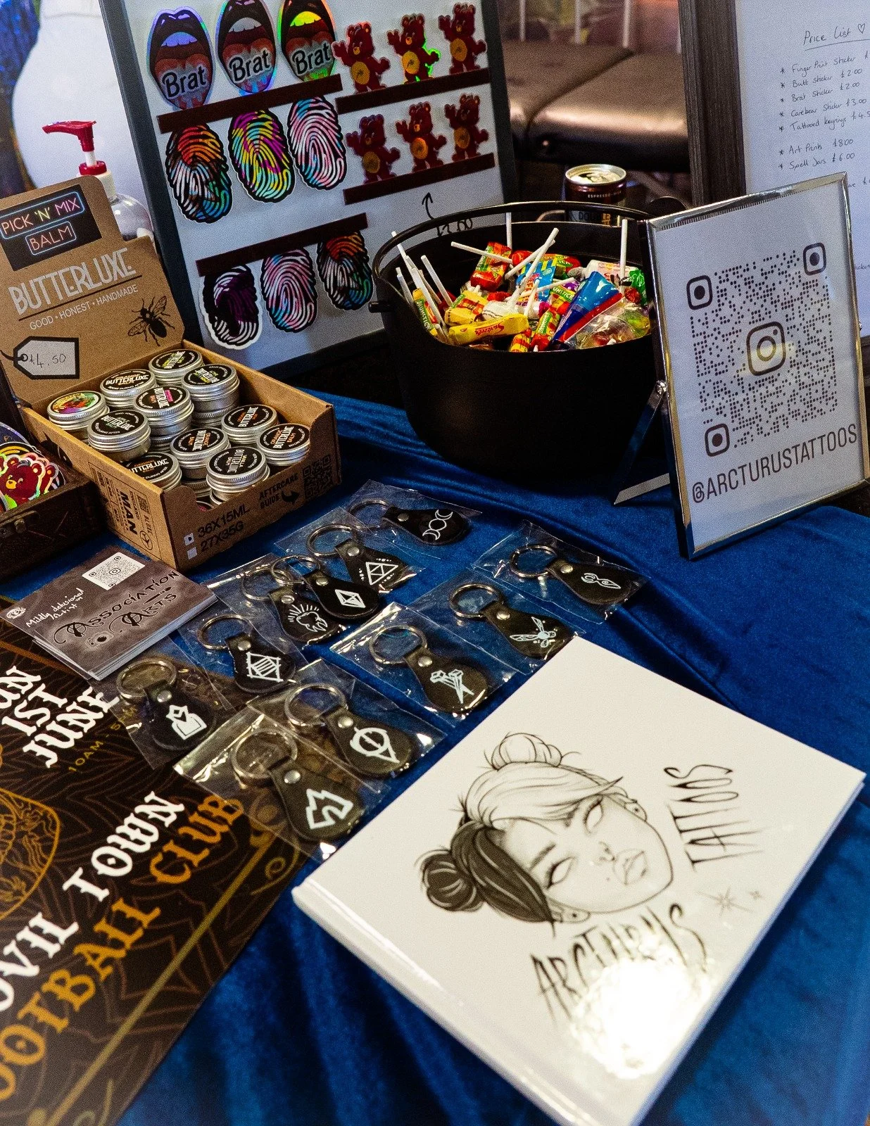 Tattoo booth display with sticker keychains, colorful candies, a digital display showing sticker designs, a QR code sign, and a sketchbook with a portrait of a woman and the words 'Arcturus Tattoos'.
