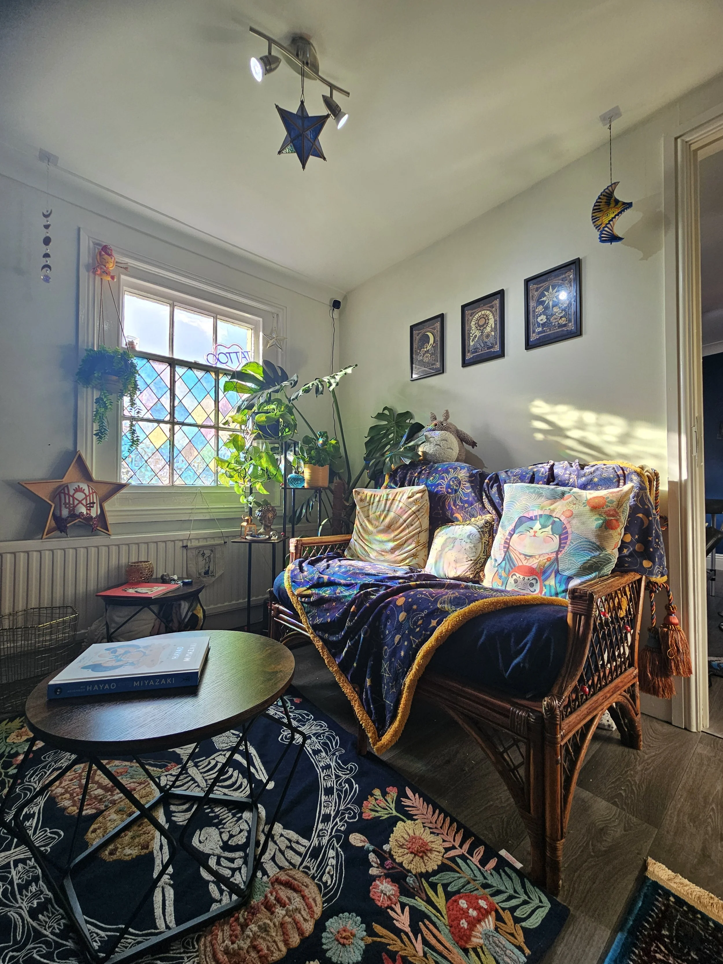 Cozy living room with a wicker couch draped in dark blue celestial-themed fabric, decorated with colorful cushions featuring cartoon designs, in front of a window with stained glass panels, surrounded by houseplants, wall art, hanging decorations, and a small wooden coffee table with a book.