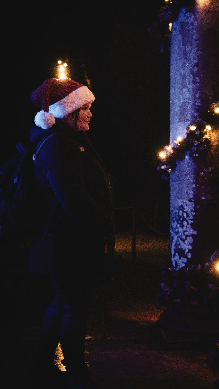 A woman wearing a Santa hat and dark winter coat stands in front of a Christmas tree decorated with lights at night.