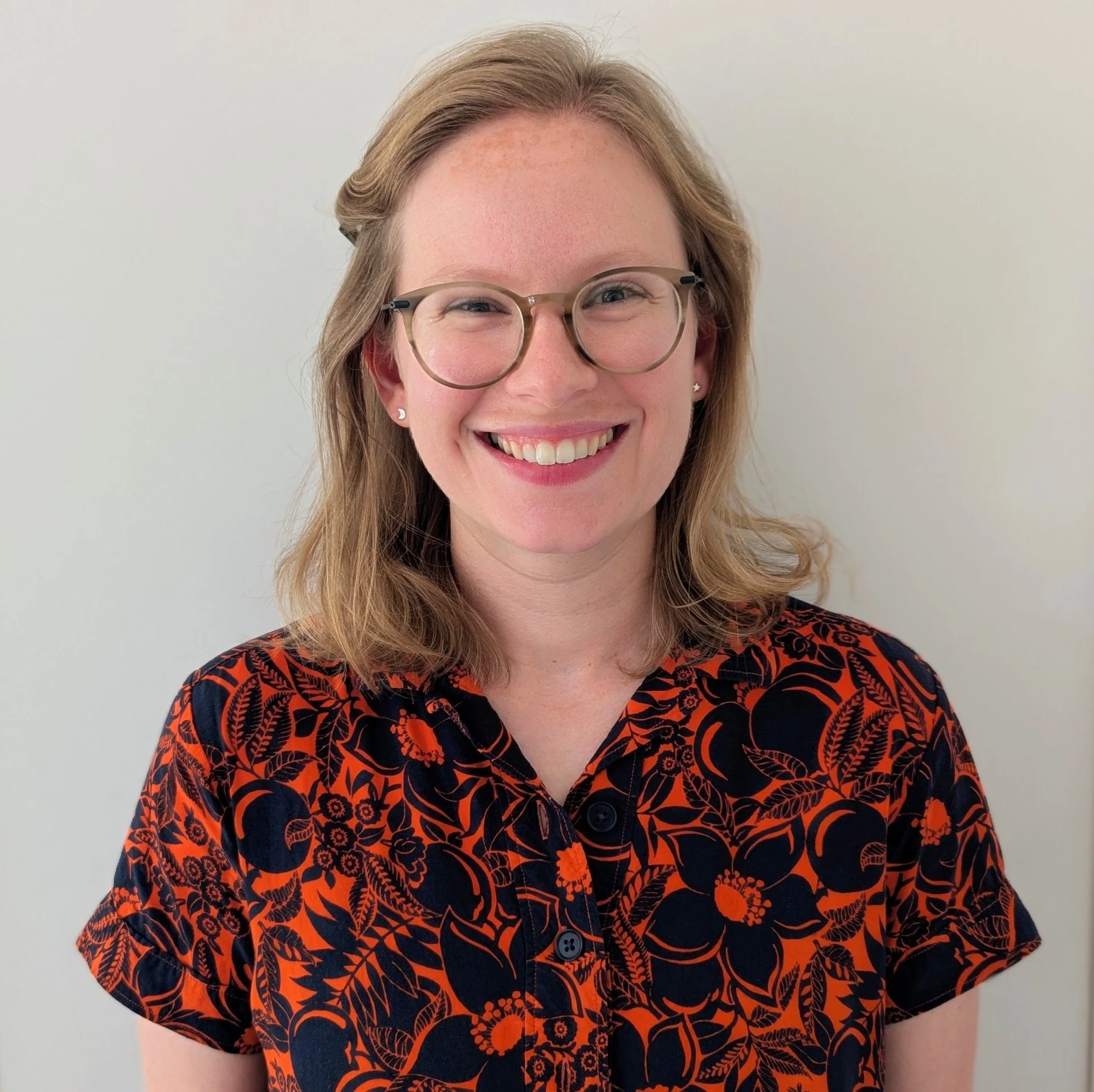 A woman with glasses and a floral-patterned shirt, smiling against a plain background.